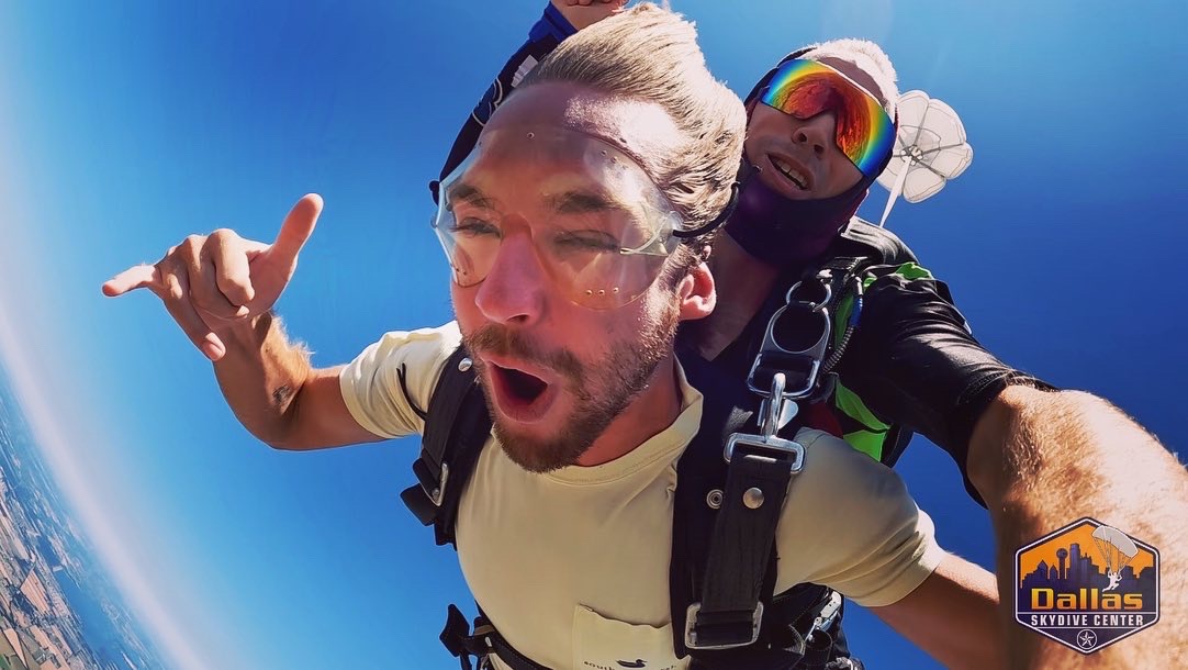 Two people skydiving with a clear blue sky background, one making a peace sign, both wearing harnesses and goggles.