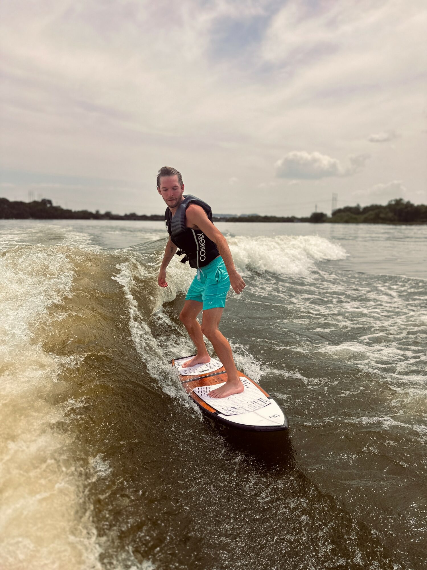Child surfing on a wave, wearing a life jacket and blue shorts, with a cloudy sky in the background.