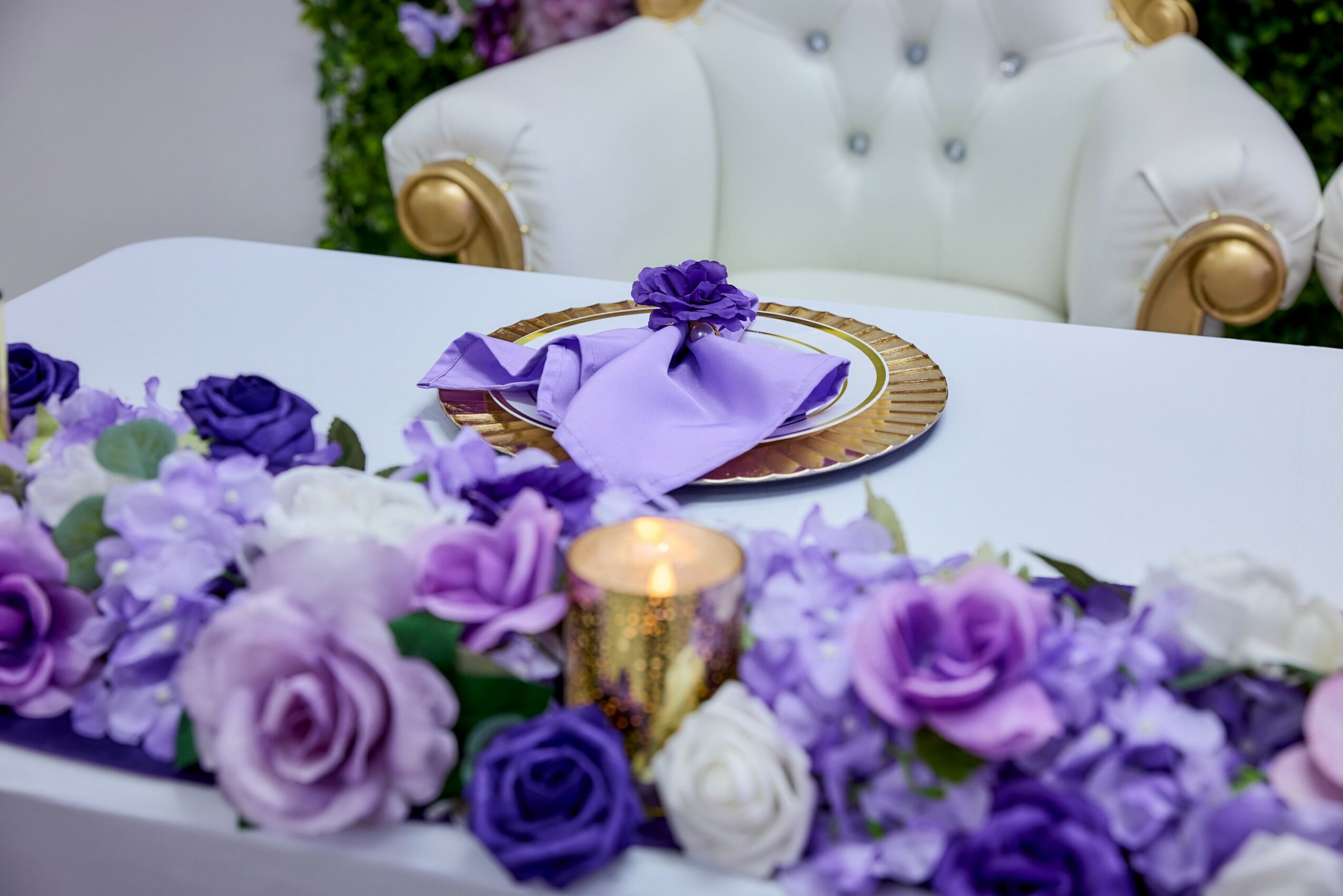 Elegant table setting with purple and white flowers, gold accents, and a white chair in the background.
