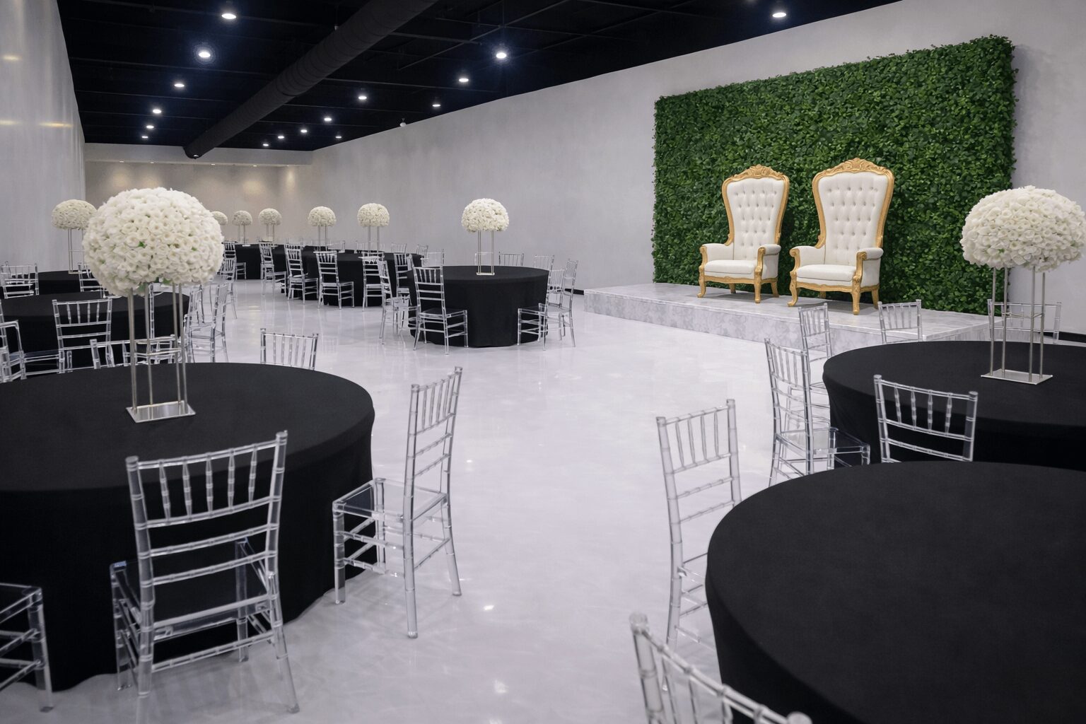 Event space with round tables, black tablecloths, white chairs, and a green wall with two chairs on a stage, 20 words.
