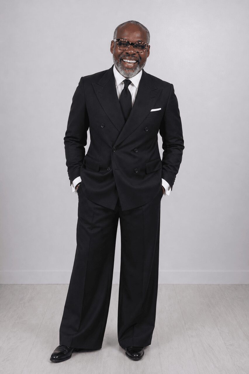 Man in a black suit, white shirt, and black tie standing with hands in pockets, smiling, against a plain light background.