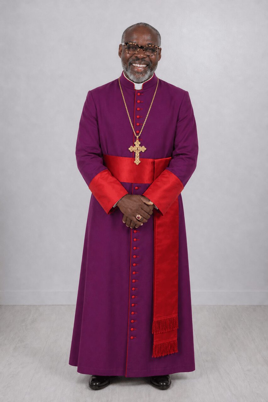 A smiling man in purple clerical attire with a gold cross necklace, standing against a plain background.