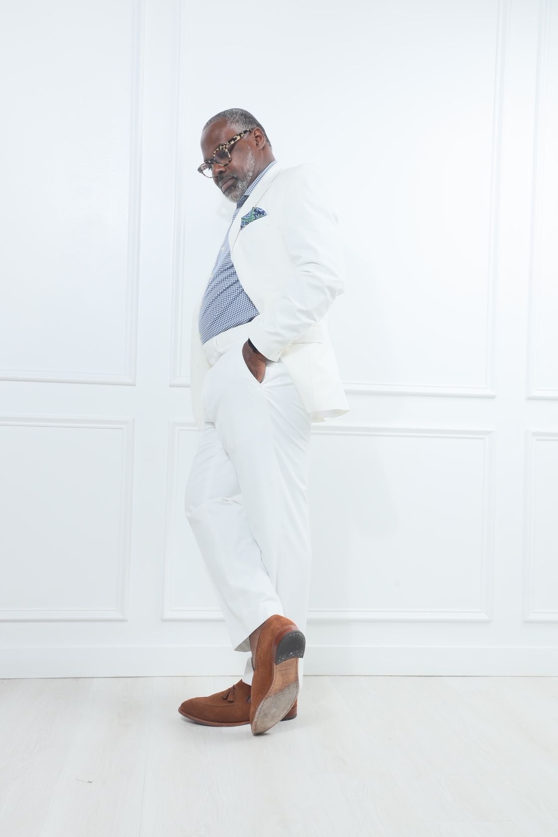 Man in white suit and brown shoes standing sideways against a white wall, with a blue bag on his shoulder.