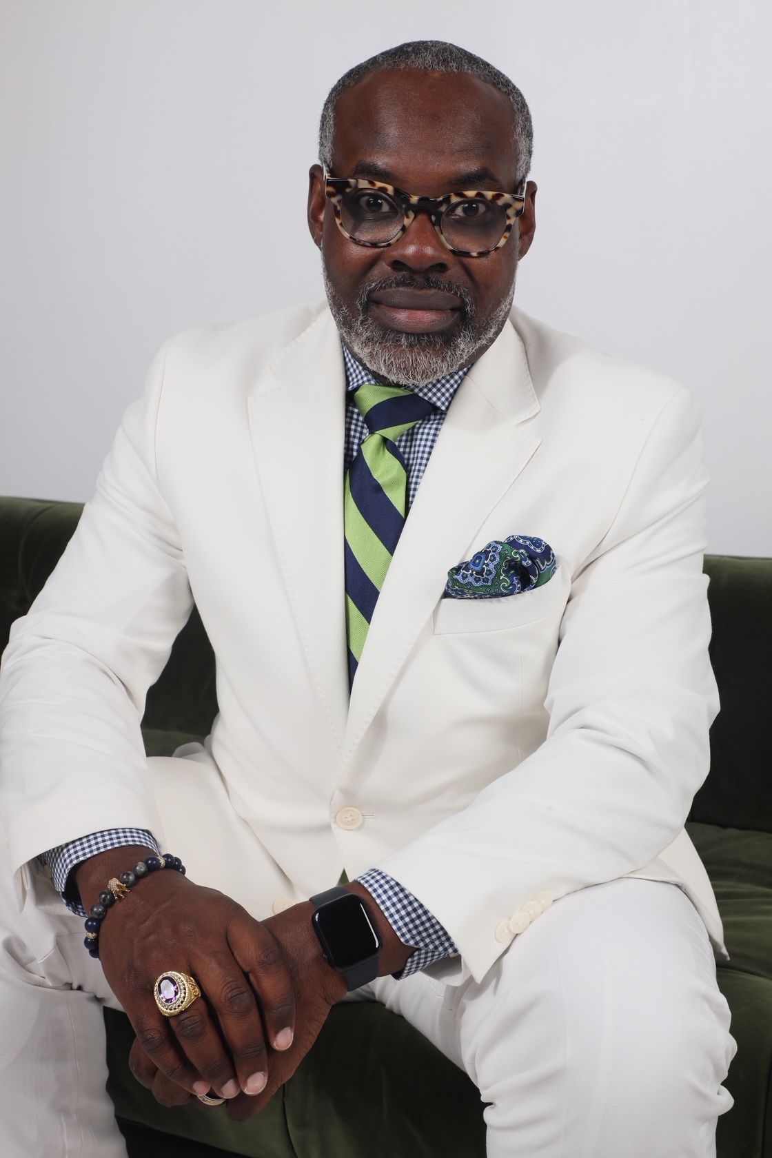 Man in white suit with glasses, sitting with hands clasped, wearing a watch and rings, against a plain background.