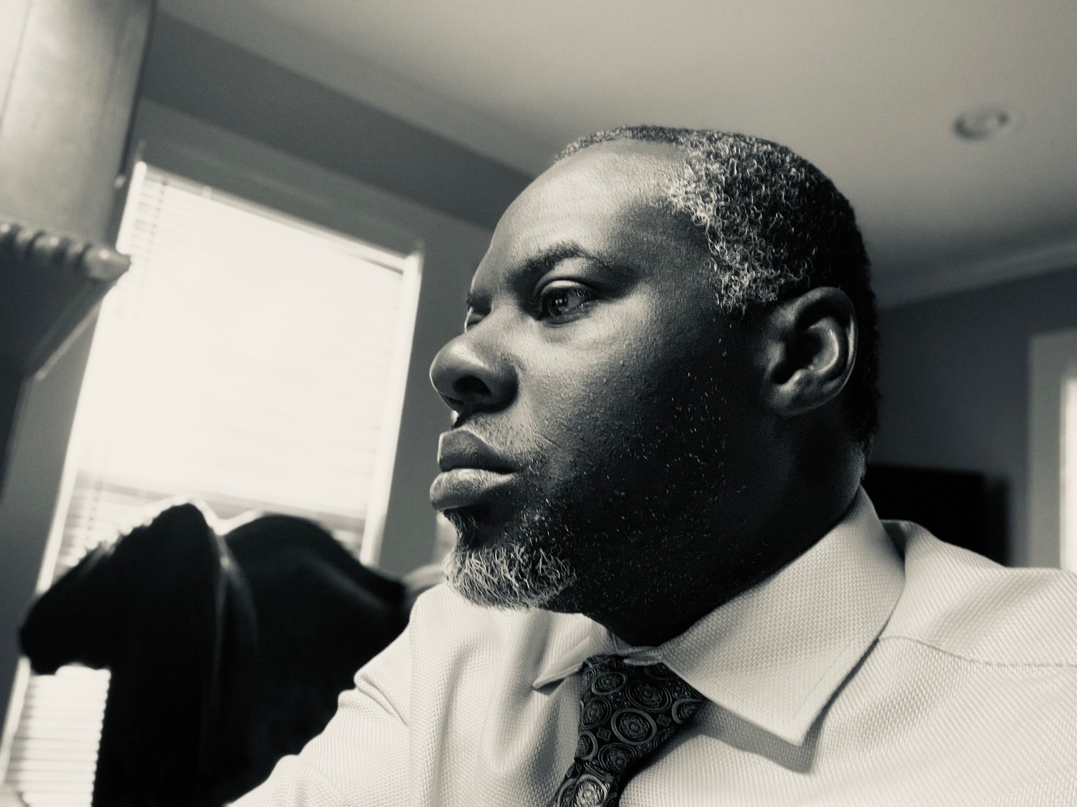Black and white portrait of a man in a shirt and tie looking out a window, with a serious expression.
