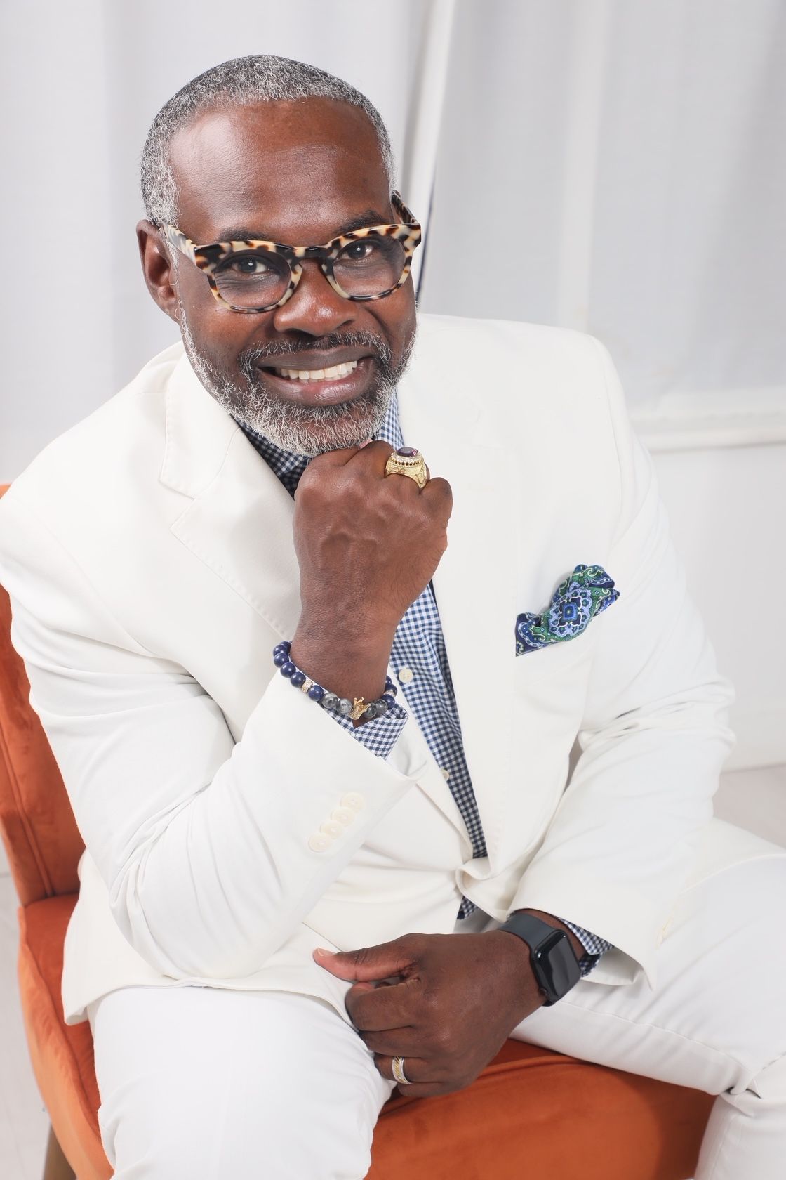 Smiling man with glasses, white shirt, and patterned pocket square, sitting on a couch, holding his chin with his hand.