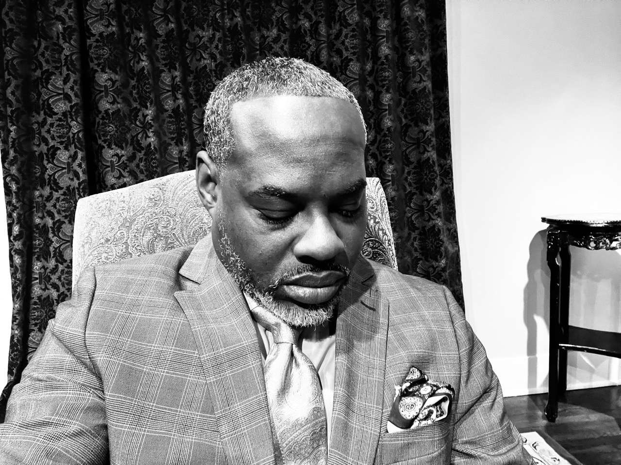 Man in suit sitting with head slightly bowed, in a room with patterned curtains and a chair in the background.