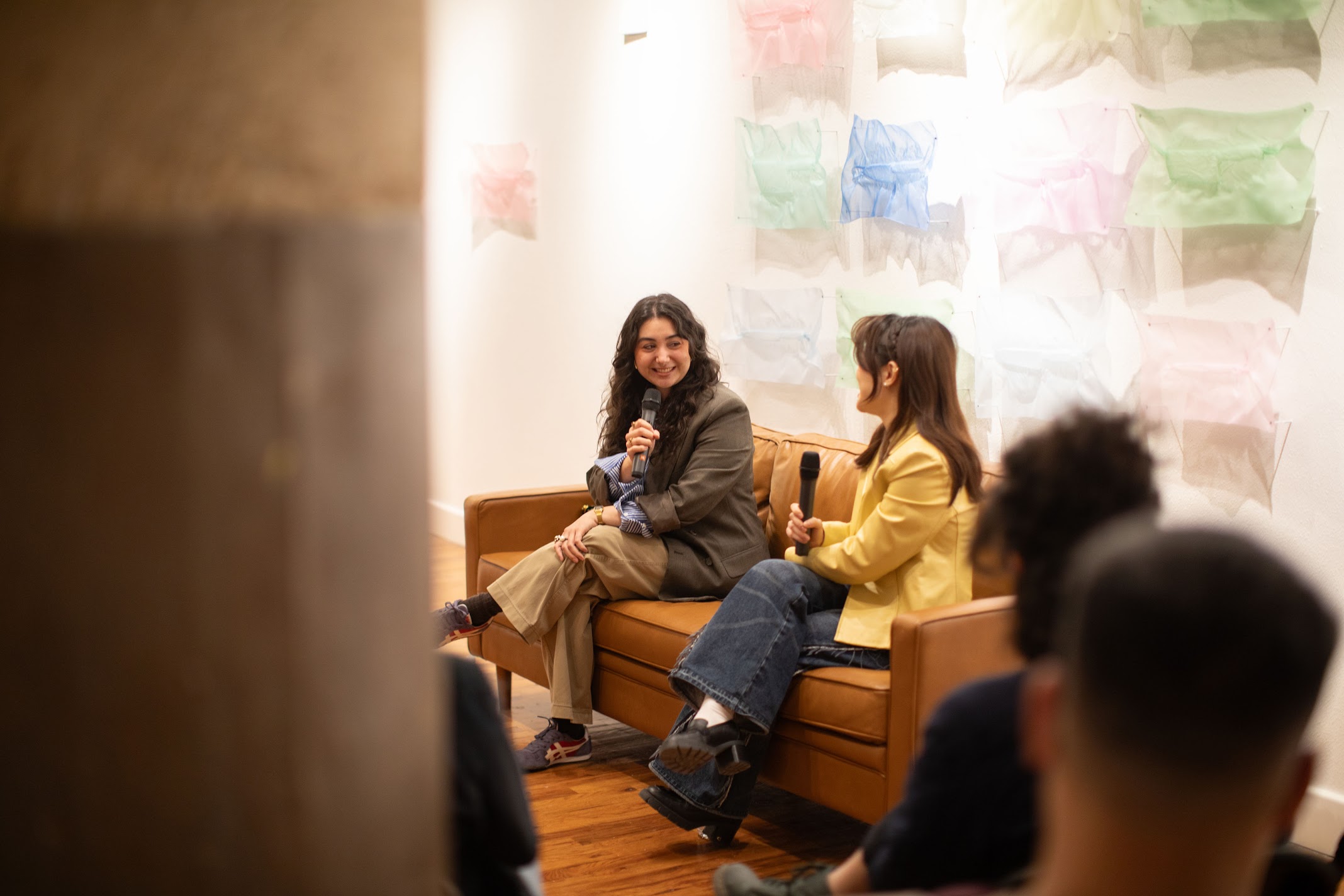 Two women sit on a sofa, engaged in conversation, with a colorful wall behind them. Several people are in the foreground.