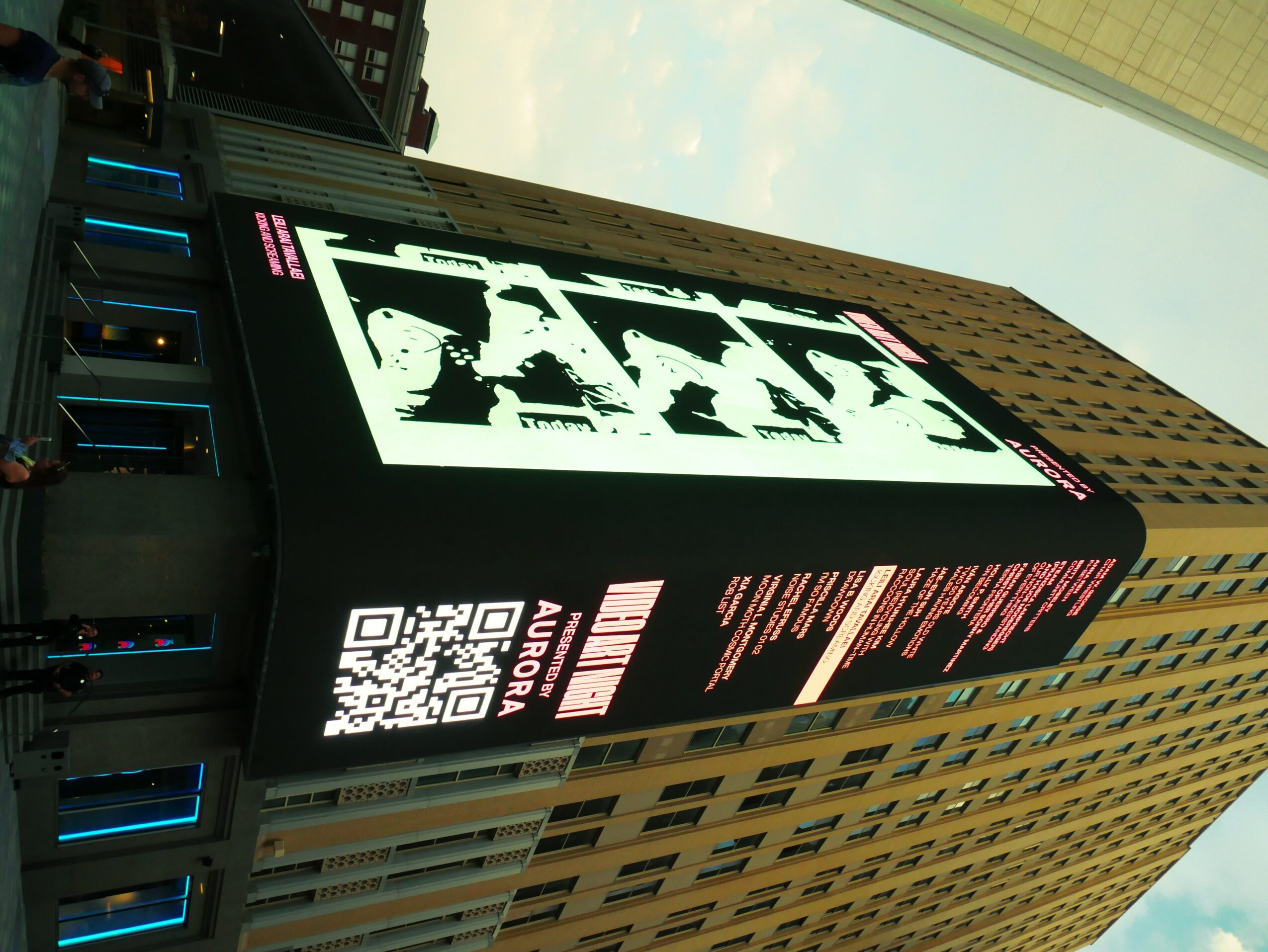 Digital billboard displaying black and white images and text, with a QR code, against a city building background.
