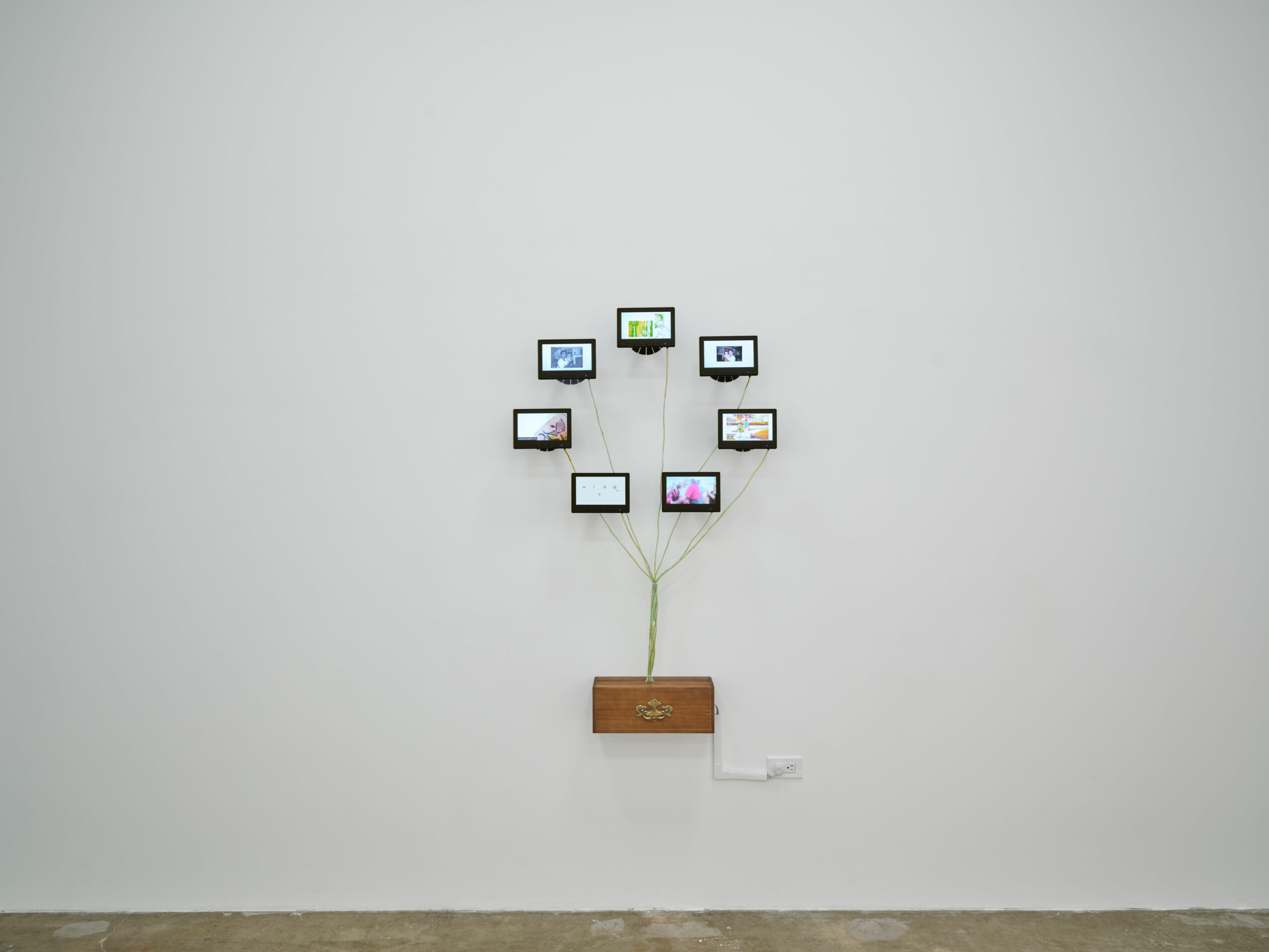 Wall with nine small framed screens arranged in a circle, connected to a wooden box below, on a plain white wall.