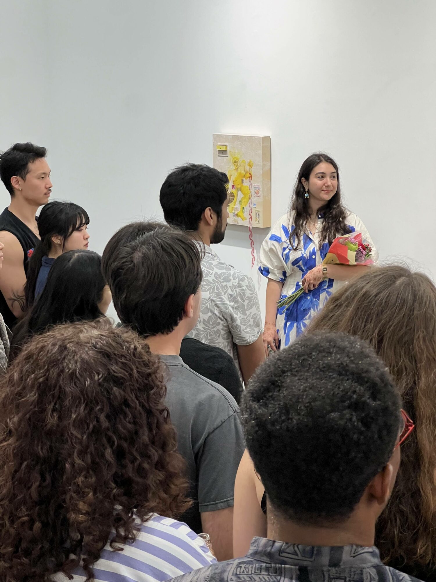 Group of people gathered indoors, with a woman holding a bouquet of flowers, standing near a wall with artwork.