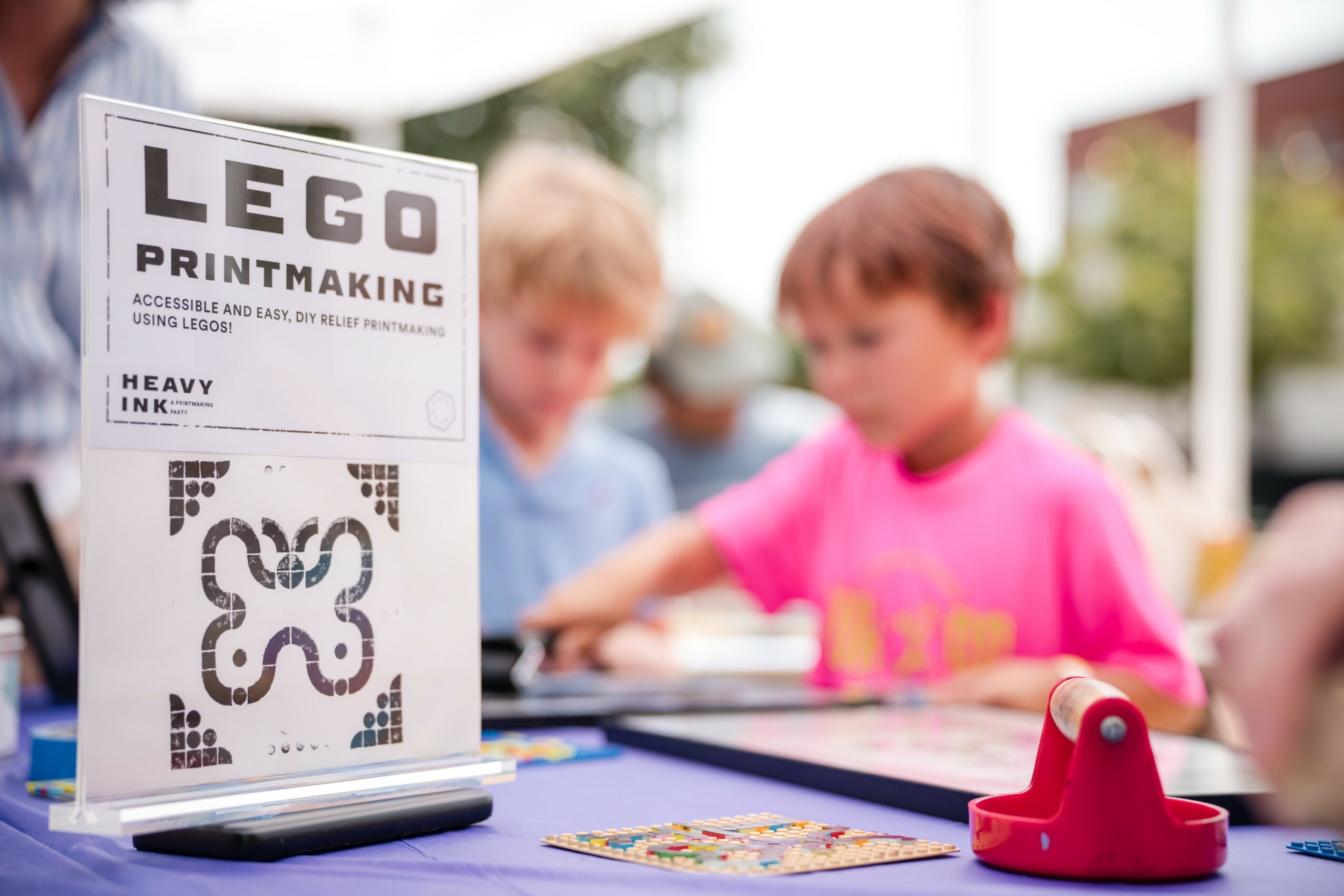 Two children playing with colorful toys outdoors, with a sign about LEGO printmaking in the foreground.