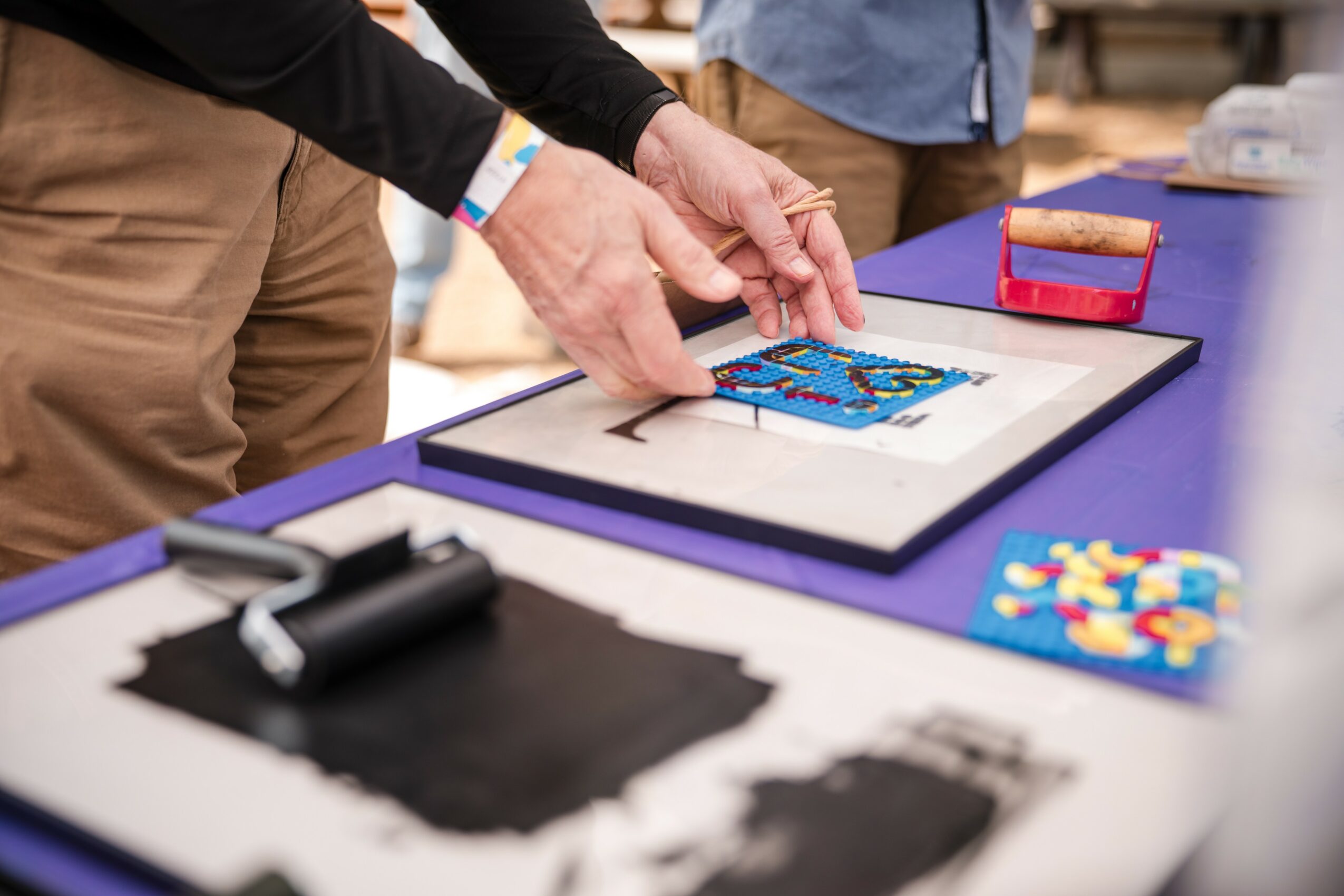 Two people work on a framed art piece on a purple table, with art supplies nearby, in a creative workspace.