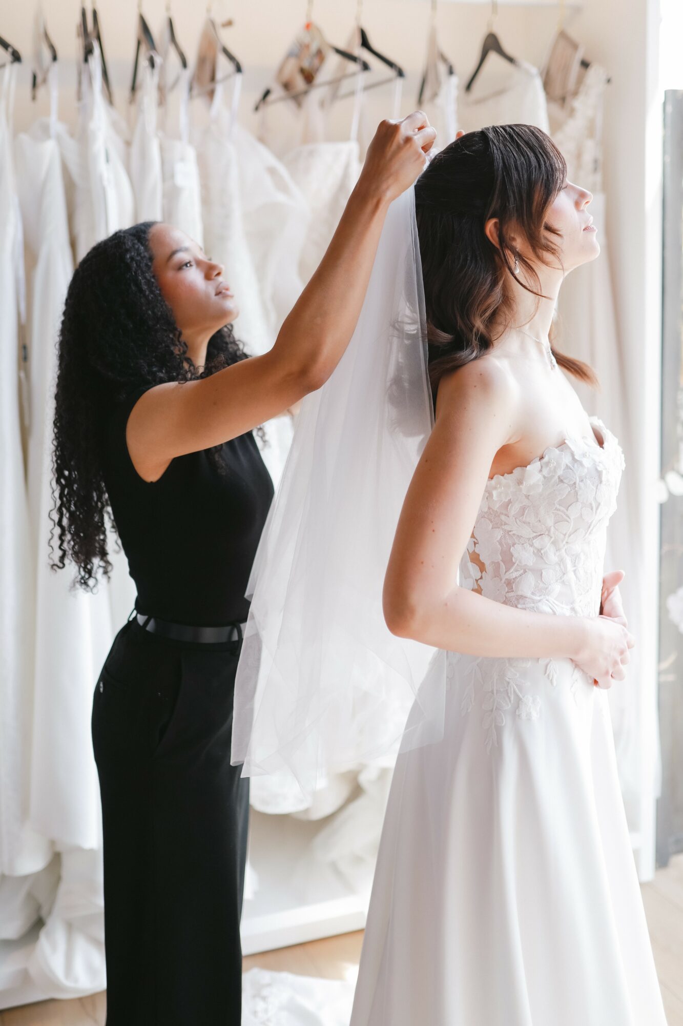 A woman helps a bride with her veil in a bridal shop, surrounded by wedding dresses on hangers.