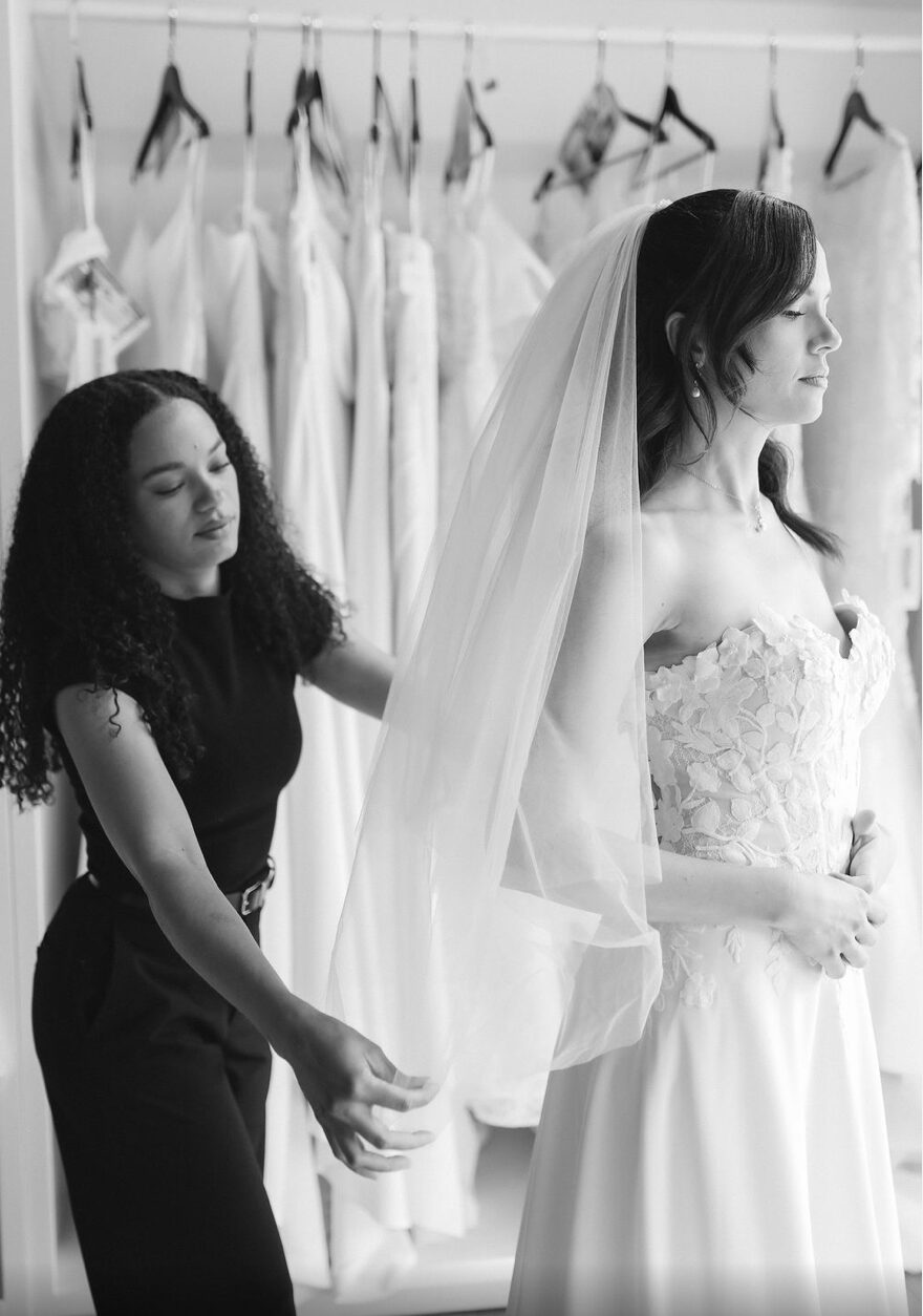 A bride in a wedding dress and veil stands with a woman adjusting her dress, in front of hanging wedding gowns.