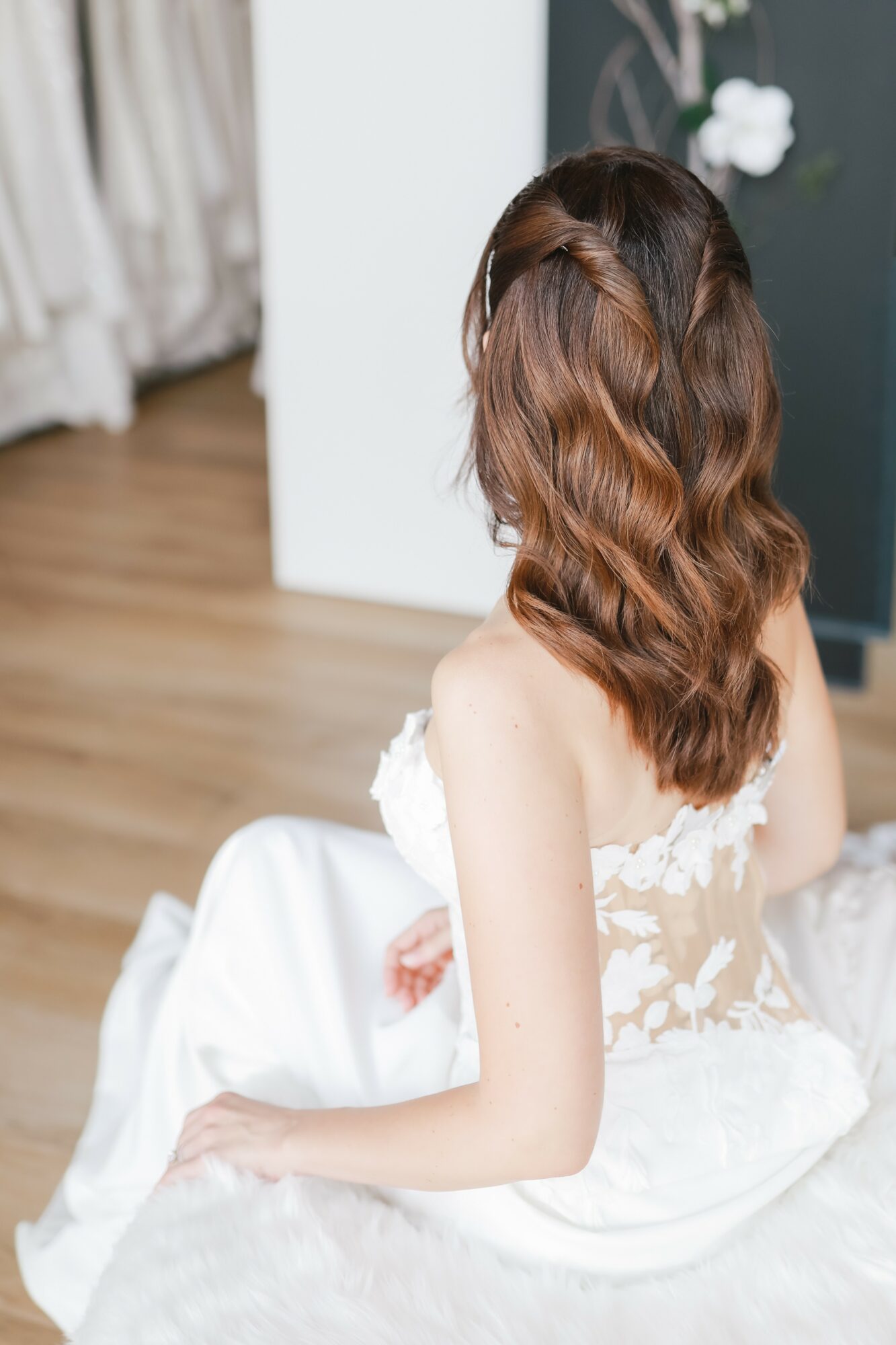 Woman with wavy brown hair sitting on a white surface, wearing a white dress, in a room with wooden floor and dark wall.