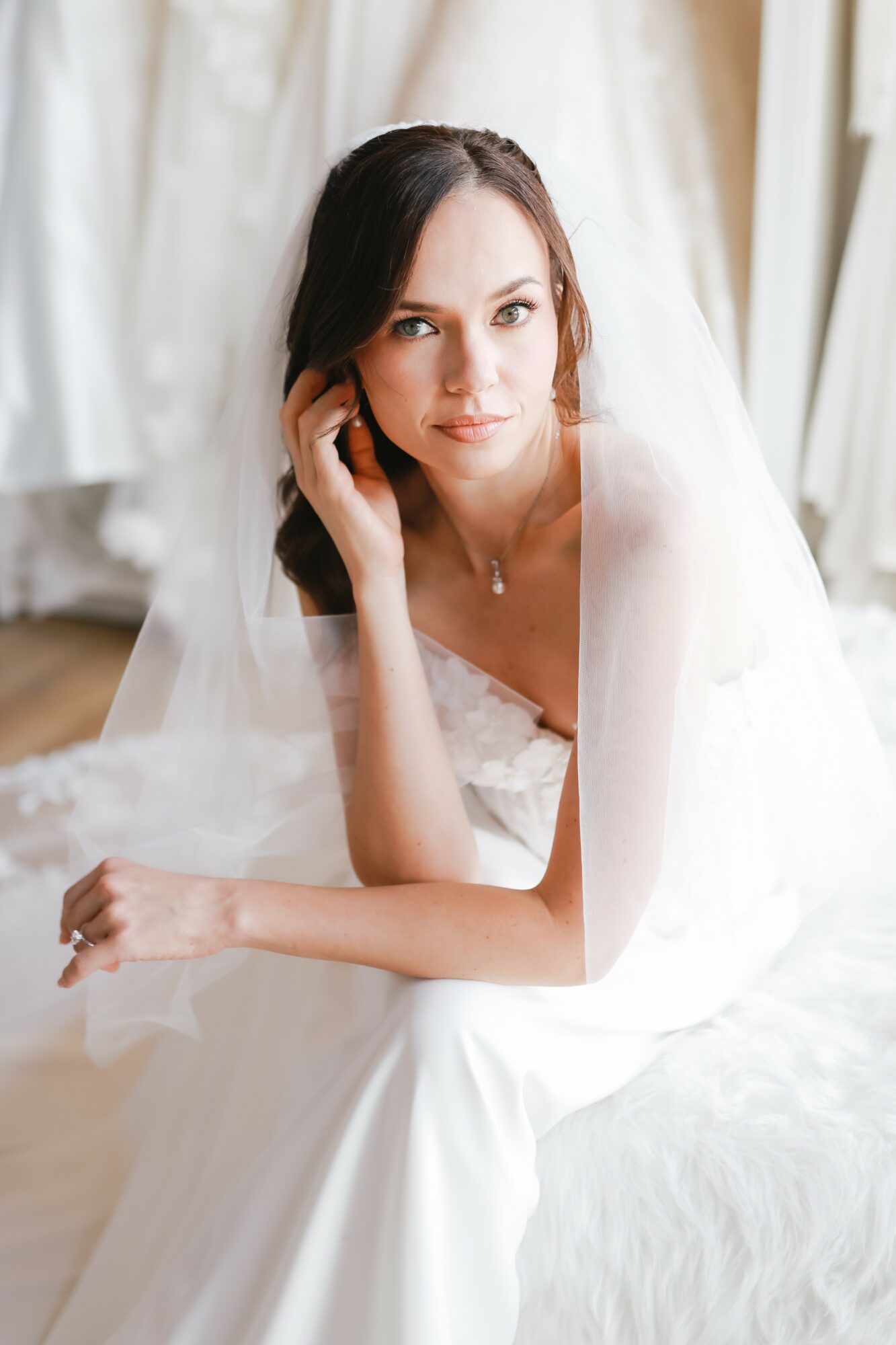 Woman in wedding dress sitting indoors, looking at camera, with veil and dark hair, surrounded by white curtains.
