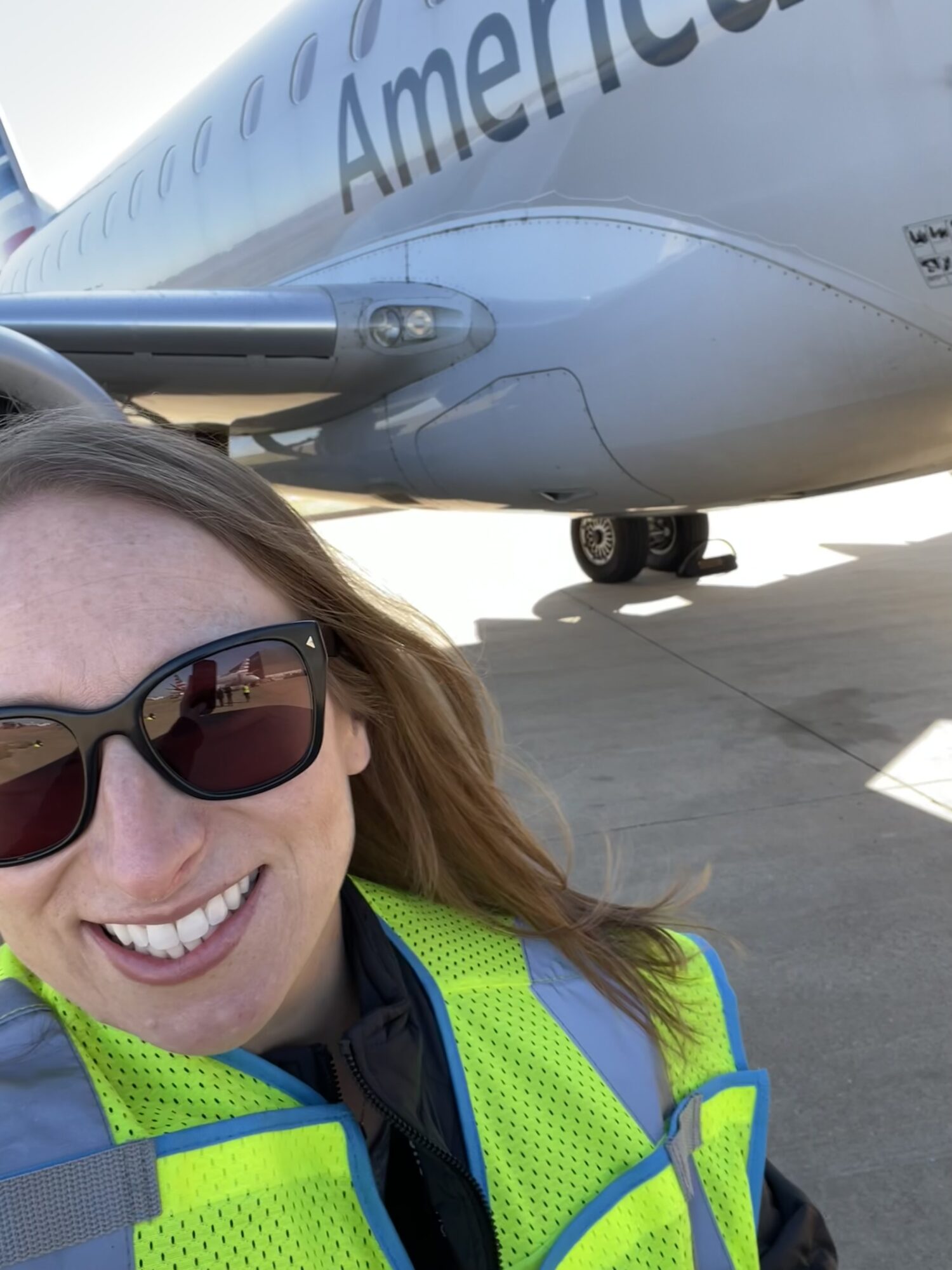 Person wearing sunglasses and a high-visibility vest smiling in front of an airplane on the tarmac.