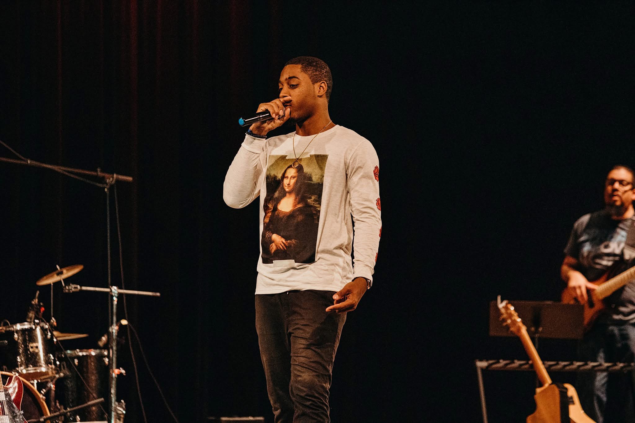 Young man singing into a microphone on stage with musical instruments in the background.