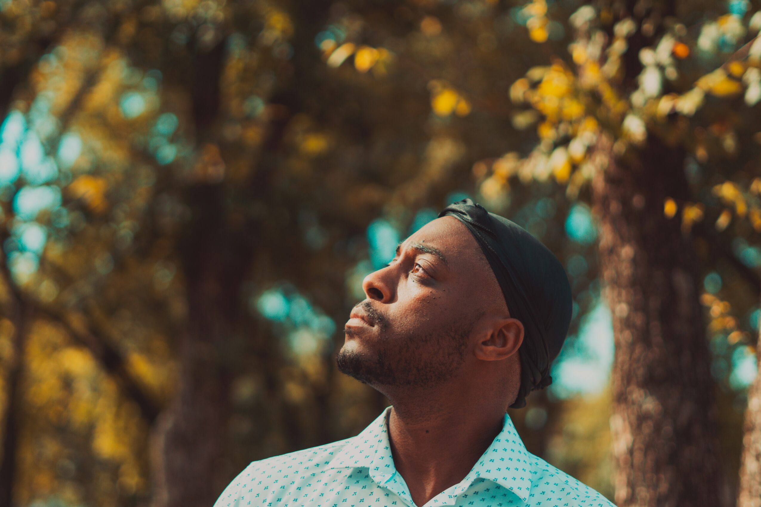 Man with closed eyes outdoors, surrounded by trees with yellow leaves, sunlight on face, wearing a white shirt and headband.