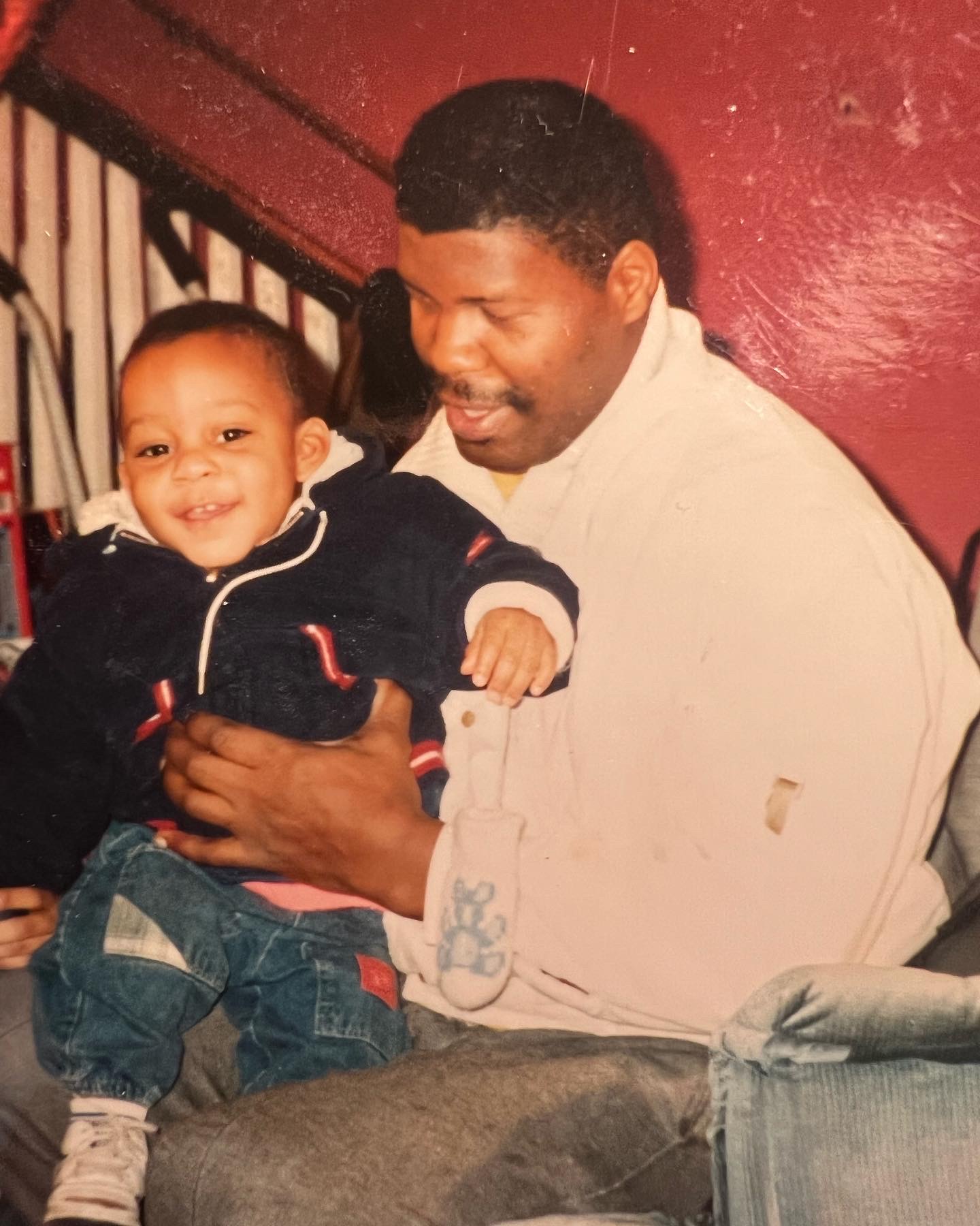 Adult man holding smiling young child, both sitting indoors against a red wall, man wearing a white shirt, child in dark jacket and jeans.