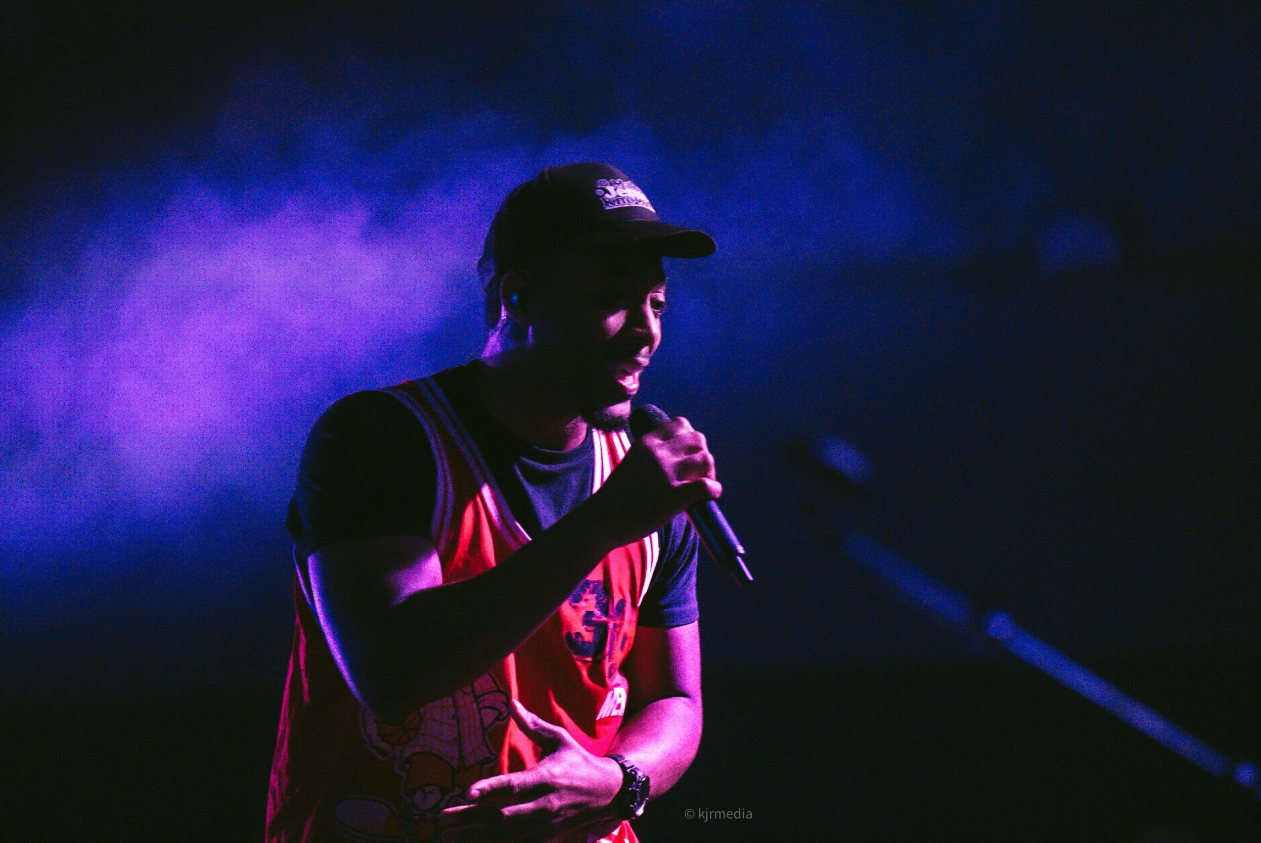 Person wearing a cap and holding a microphone on stage with purple and blue lighting, facing left.
