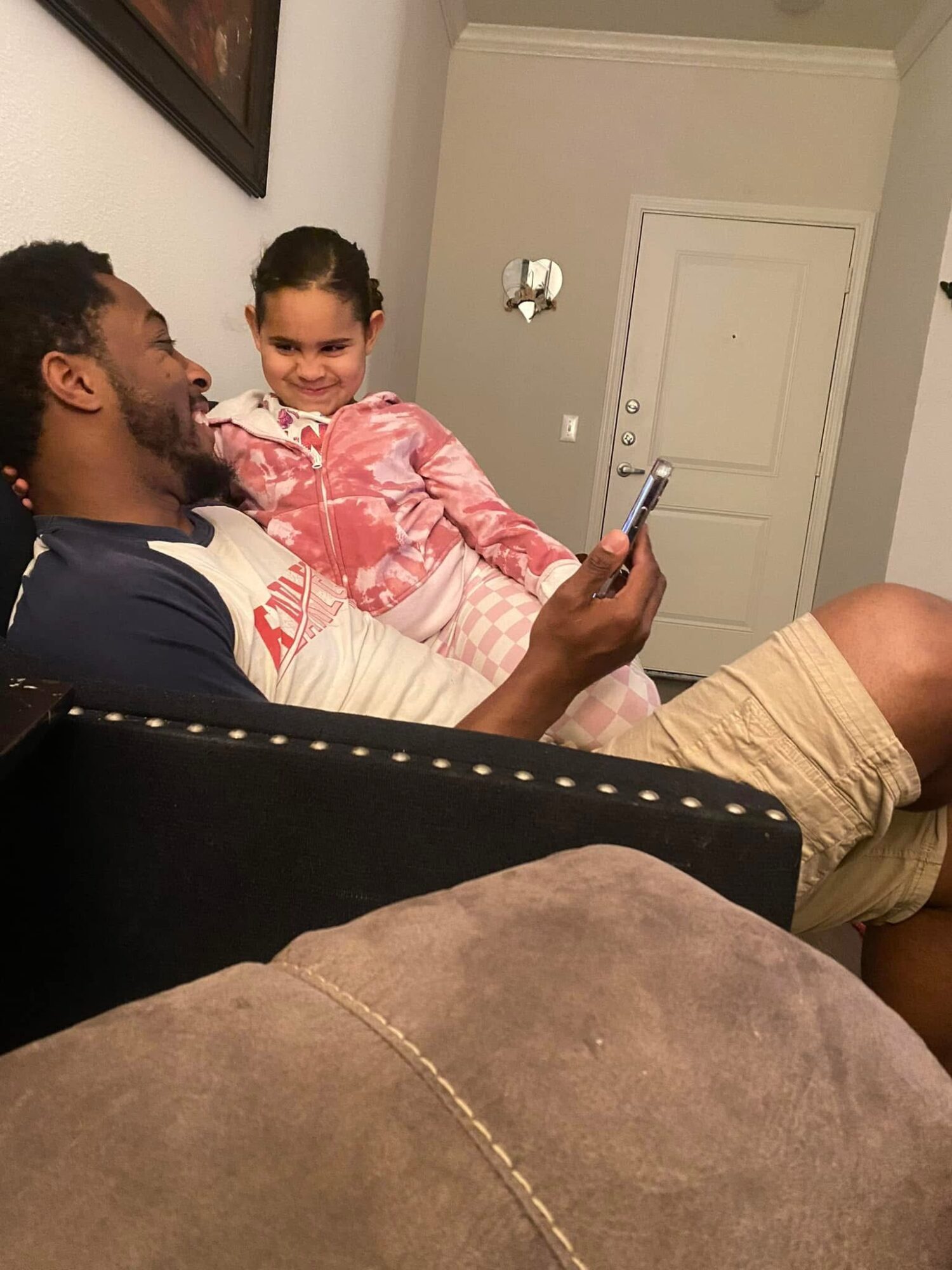 Man and young girl sitting on a couch, smiling and looking at a phone, in a room with a closed door in the background.