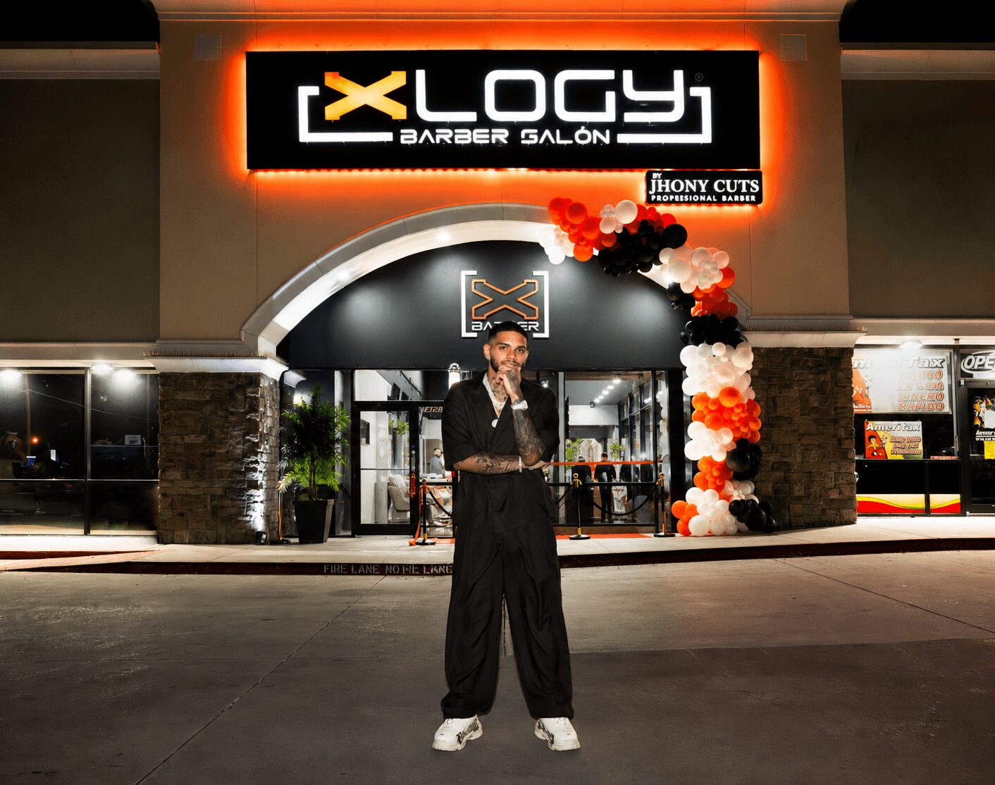 Person standing outside a barbershop with a sign reading 'XLOGY BARBER SALON' and decorative balloons, at night.