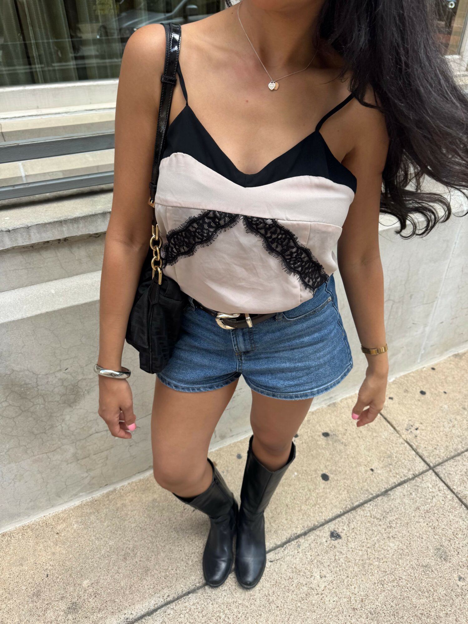 Person standing on sidewalk wearing a black top, denim shorts, and black boots, with a purse and jewelry, outdoors.