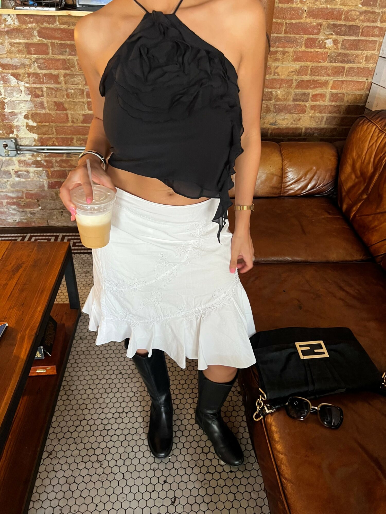 Person sitting on a couch holding a drink, wearing a black top and white skirt, with a black bag nearby.