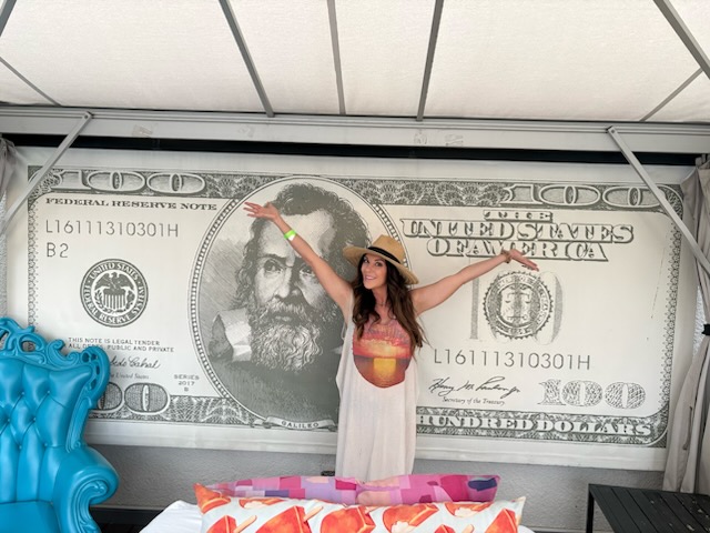Woman in a white dress and hat stands in front of a large dollar bill backdrop, pointing at the bill's face.