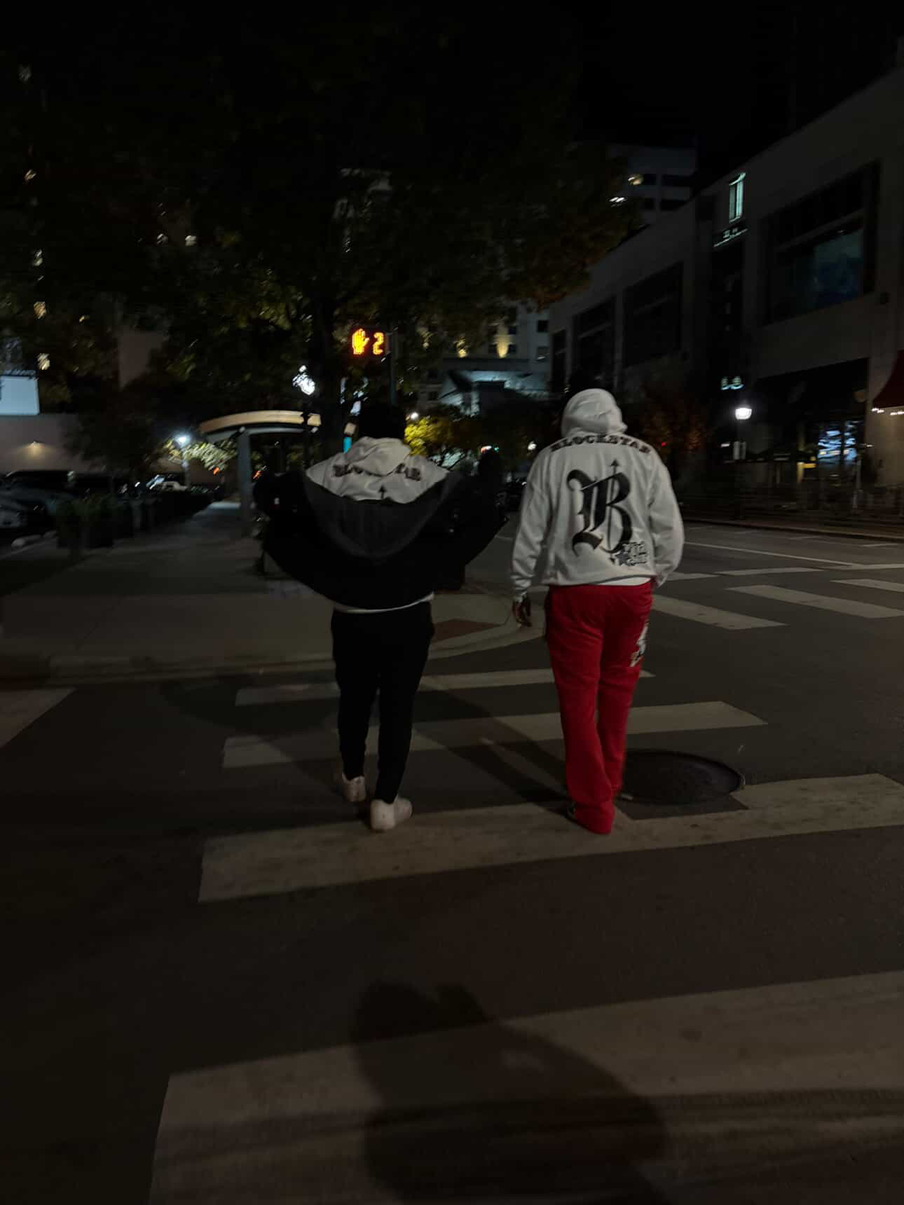 Two people walk across a street at night, one in a white hoodie and red pants, the other in dark clothing.