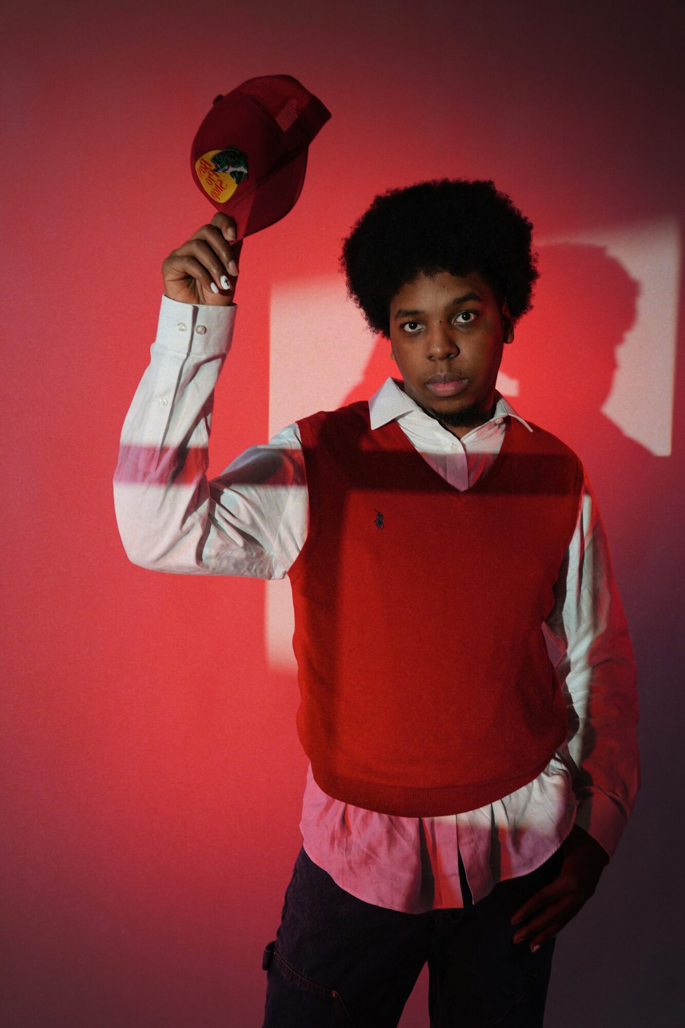 Young person with short curly hair holding a red hat, wearing a white shirt and red vest, standing against a red background.