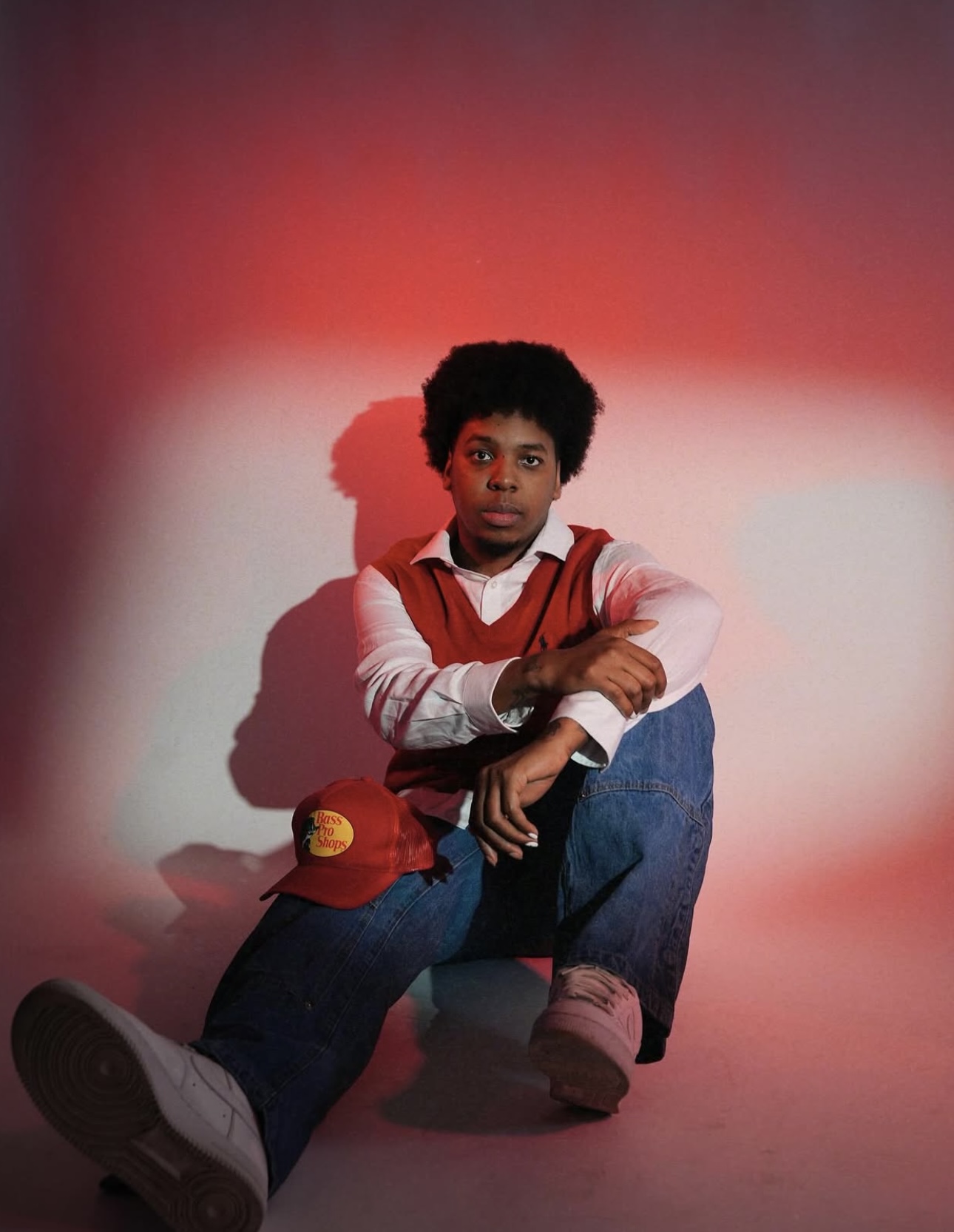 Child sitting on the floor with a red hat, wearing a red vest and jeans, against a red and white background.