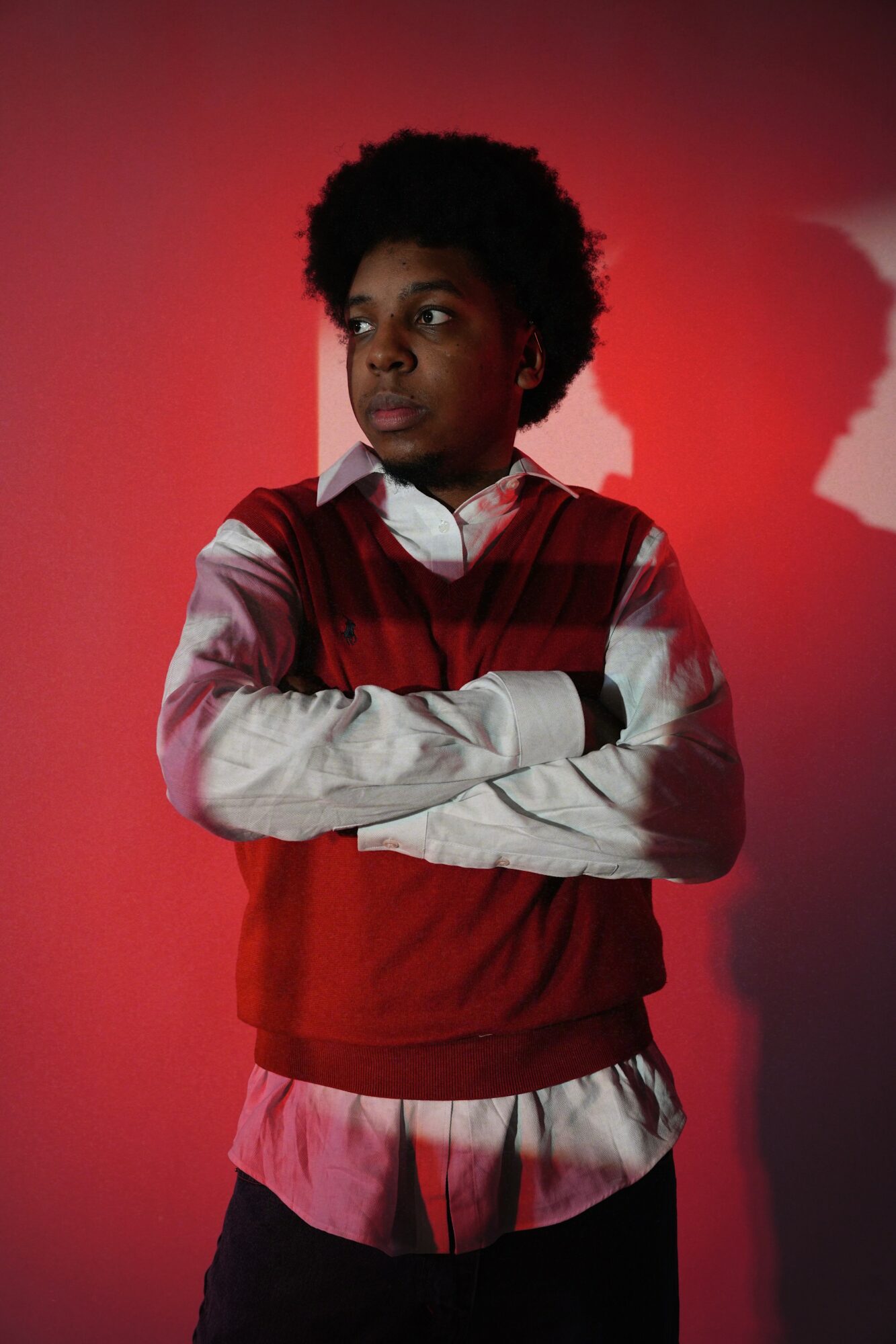 Young person with arms crossed, wearing a red vest over a white shirt, standing against a red background.