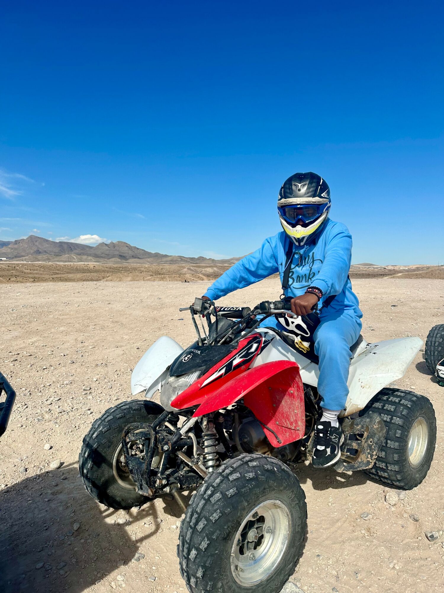 Person wearing a helmet and blue jacket riding a red and white ATV in a desert landscape under a clear blue sky.