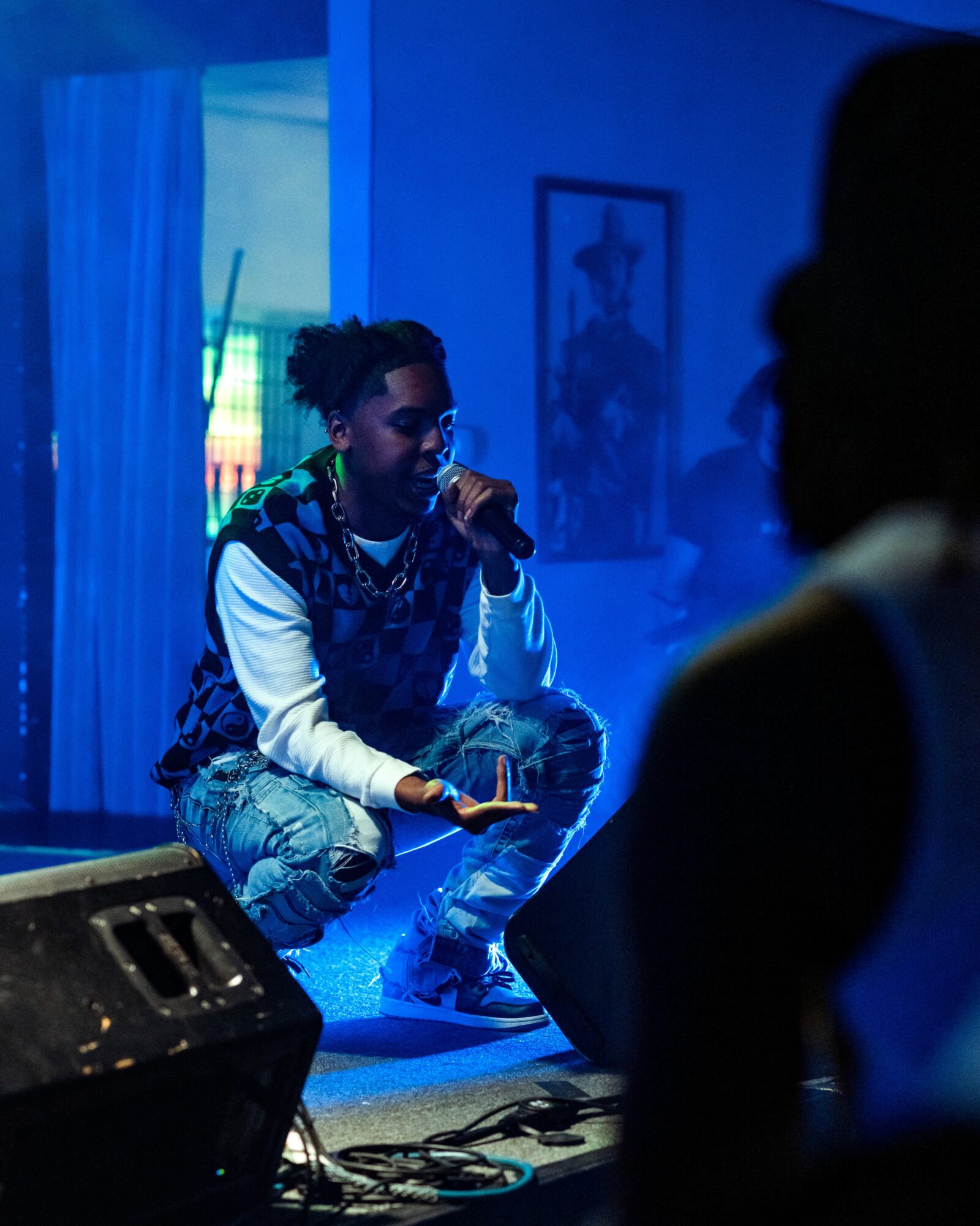 Person with short hair and patterned clothing holding a microphone, sitting on stage with blue lighting, audience in foreground.