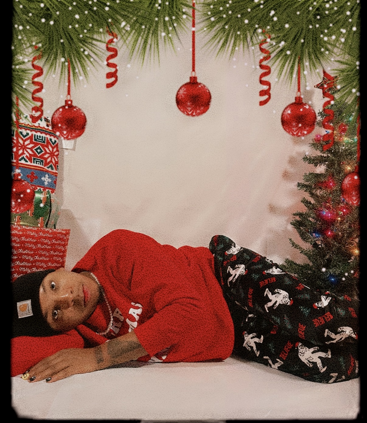 Child lying on a bed with Christmas decorations, including ornaments and a small decorated tree, above him. He wears a red shirt and patterned pants.