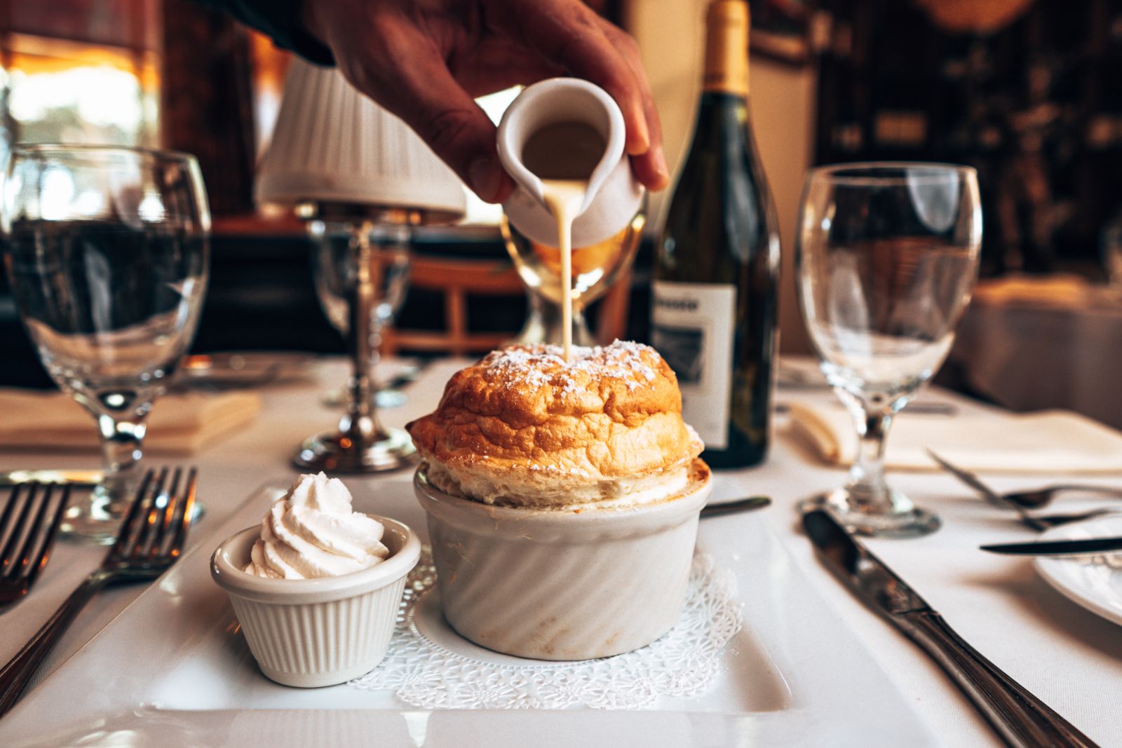 Pouring sauce over a baked dish at a restaurant table with wine glasses and utensils, with a small dessert on the side.