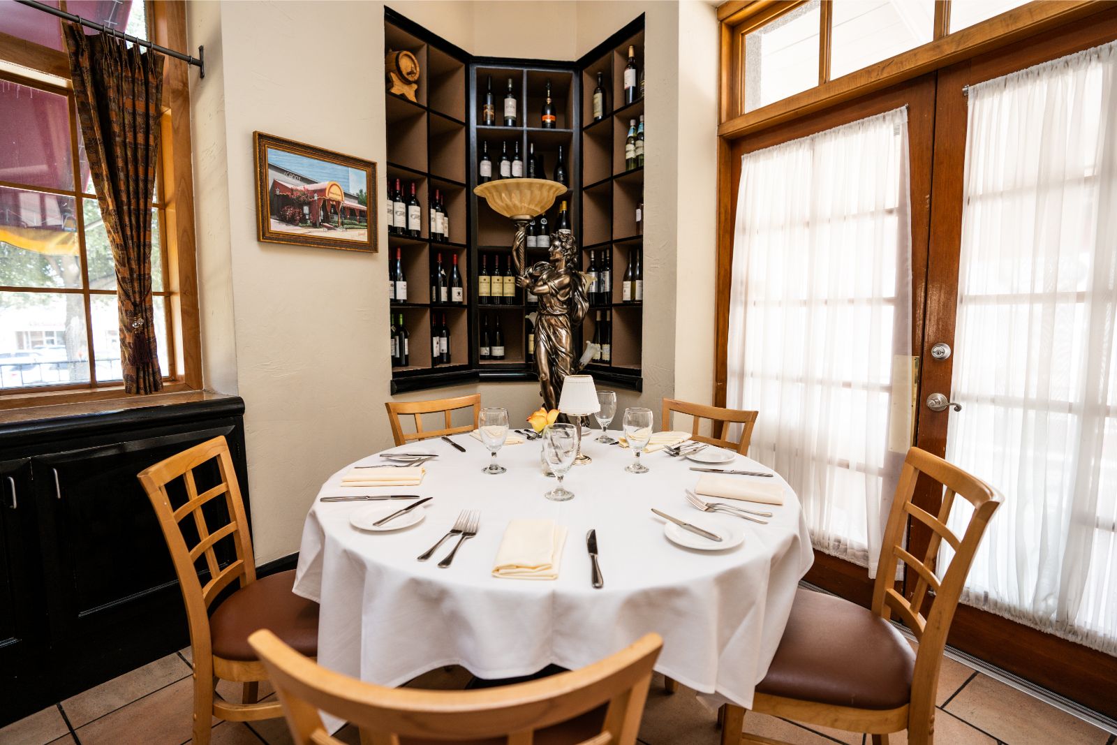 Round dining table with white tablecloth, set with plates, glasses, and silverware, surrounded by four wooden chairs, in a bright room.