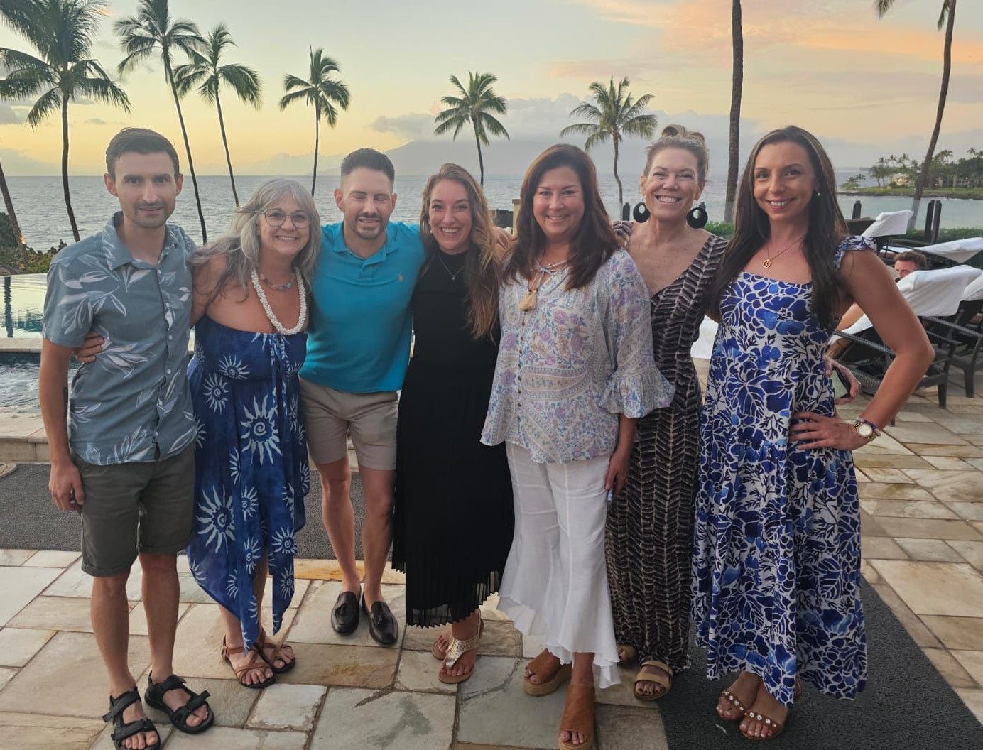 Group of seven people standing outdoors near water with palm trees, sunset sky, and parked cars in background.