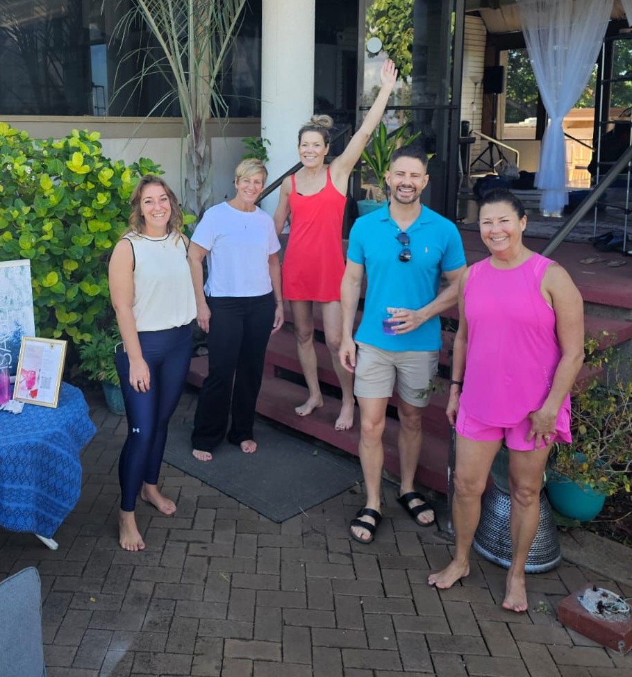 Group of five people smiling outdoors, some barefoot, standing on brick pavement near a building with plants and curtains.