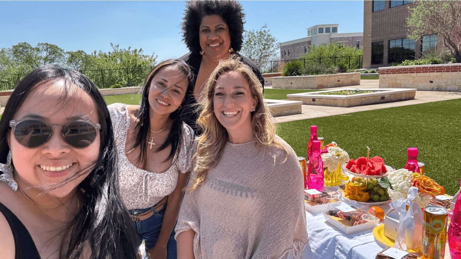 Four women smiling outdoors near a table with food and drinks, sunny day, greenery and buildings in background.