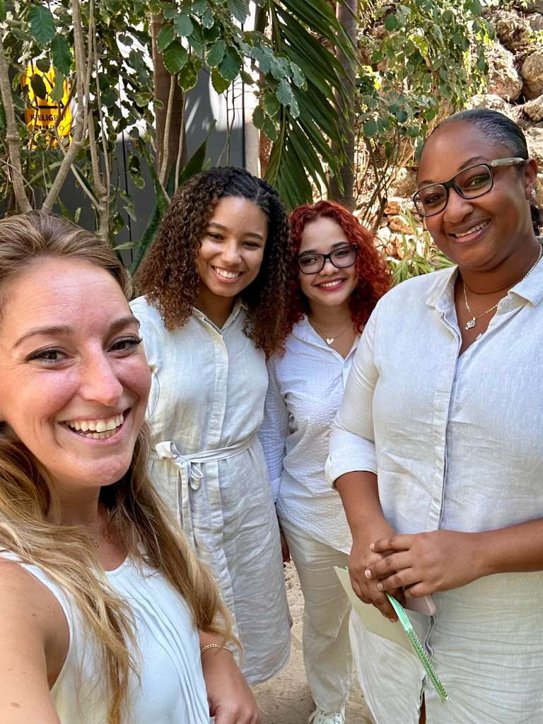 Four women smiling outdoors among greenery, wearing light-colored clothing, with one woman holding a green straw.