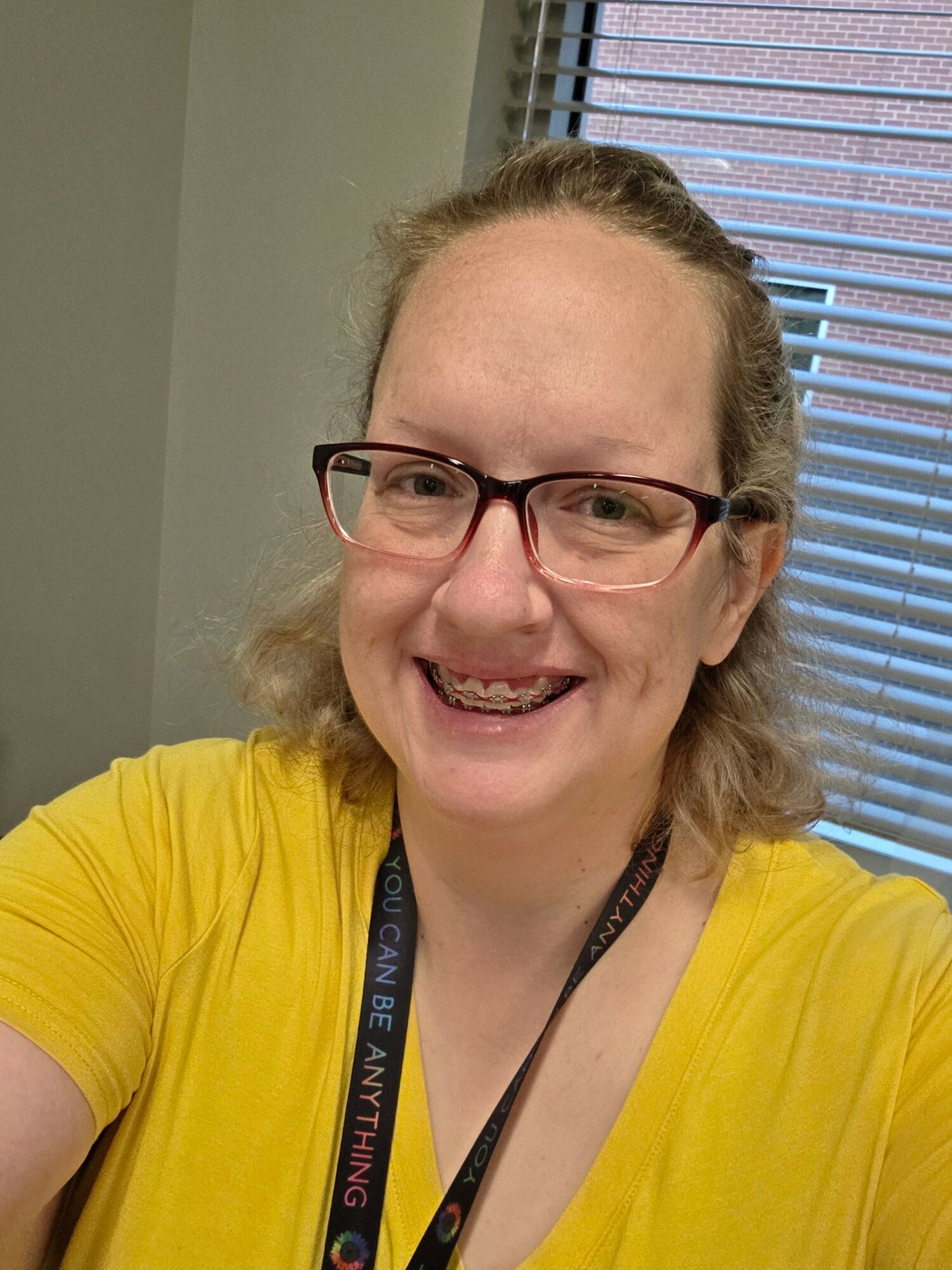Woman with glasses smiling, wearing a yellow shirt, in front of window with blinds