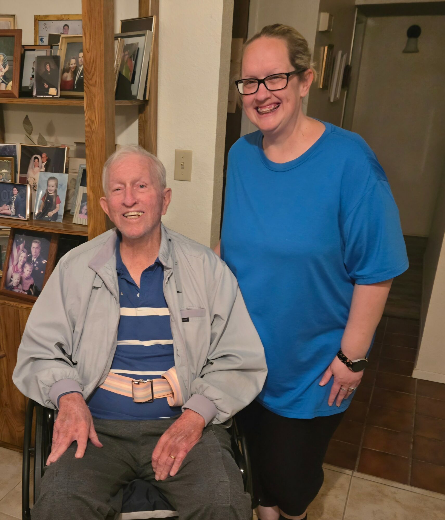 Two women smiling indoors, one seated and one standing, in front of a bookshelf with framed photos.