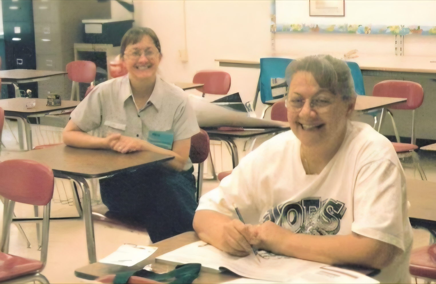 Two smiling people sitting at desks in a classroom, one adult and one child, with classroom furniture and supplies visible.