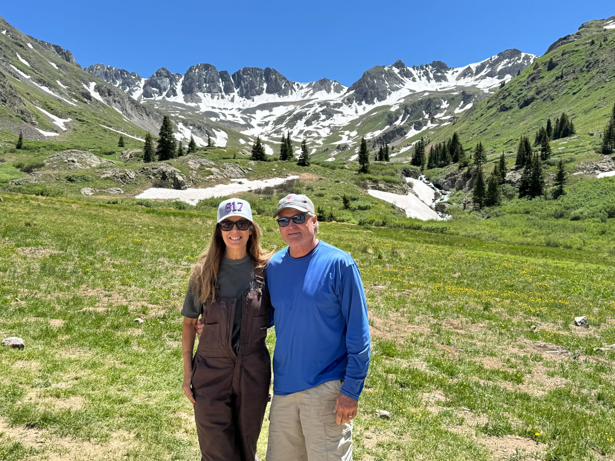 A man and woman stand together in a green field with mountains and snow in the background, sunny sky, both wearing sunglasses.