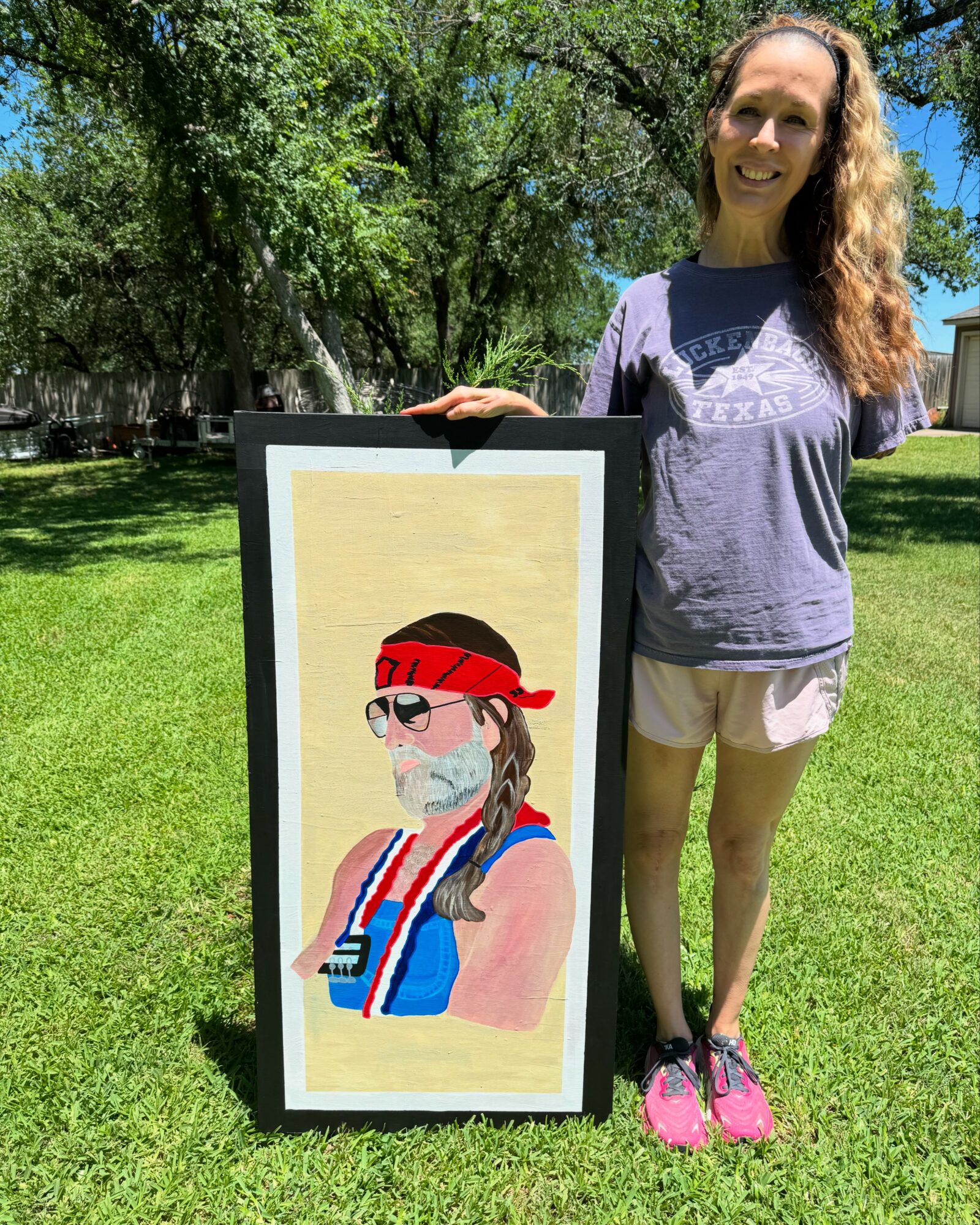 Woman standing outdoors on grass, smiling, holding a framed colorful illustration of a person with a red bandana, sunglasses, and a striped scarf.