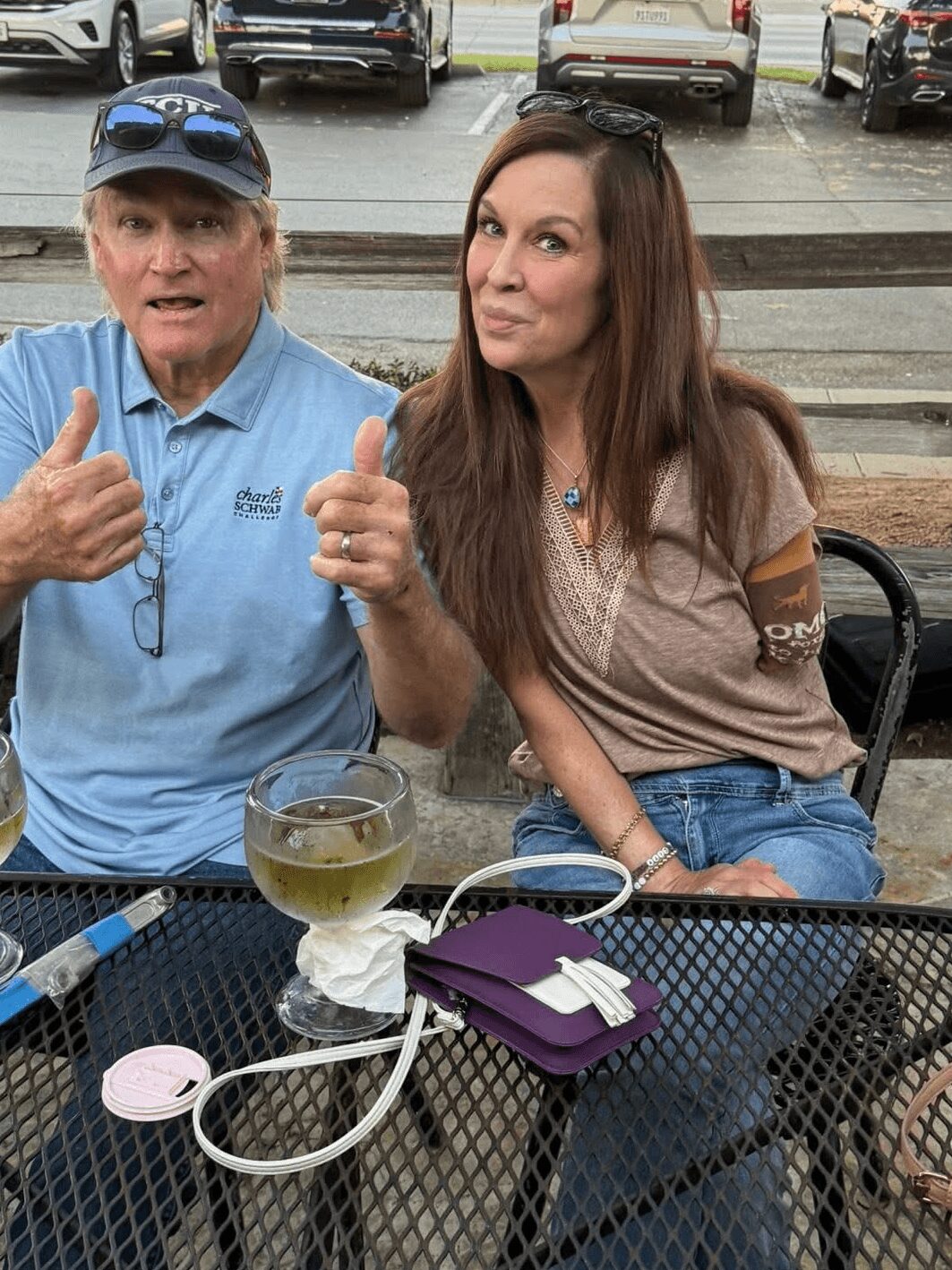 Two women sitting at an outdoor table, one gesturing with thumbs up, with cars parked in the background.